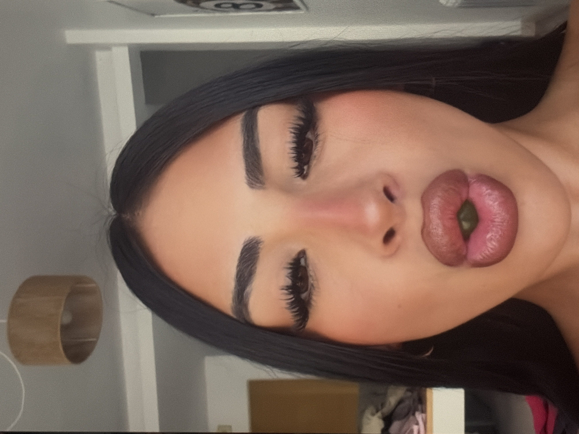 The media file is a close-up, slightly angled, portrait of a person. The person has dark hair and is wearing makeup, including pronounced eyebrows, eyelashes, and lipstick. Their lips are pursed, and a small, green object is visible within them. The background is out of focus, but appears to be an indoor setting with a shelf or wall structure and a cylindrical object that might be a lamp or a speaker. The lighting suggests an indoor environment, possibly during the day or with artificial lighting. No text is visible in the image. The overall mood is ambiguous due to the pose and the obscured face.