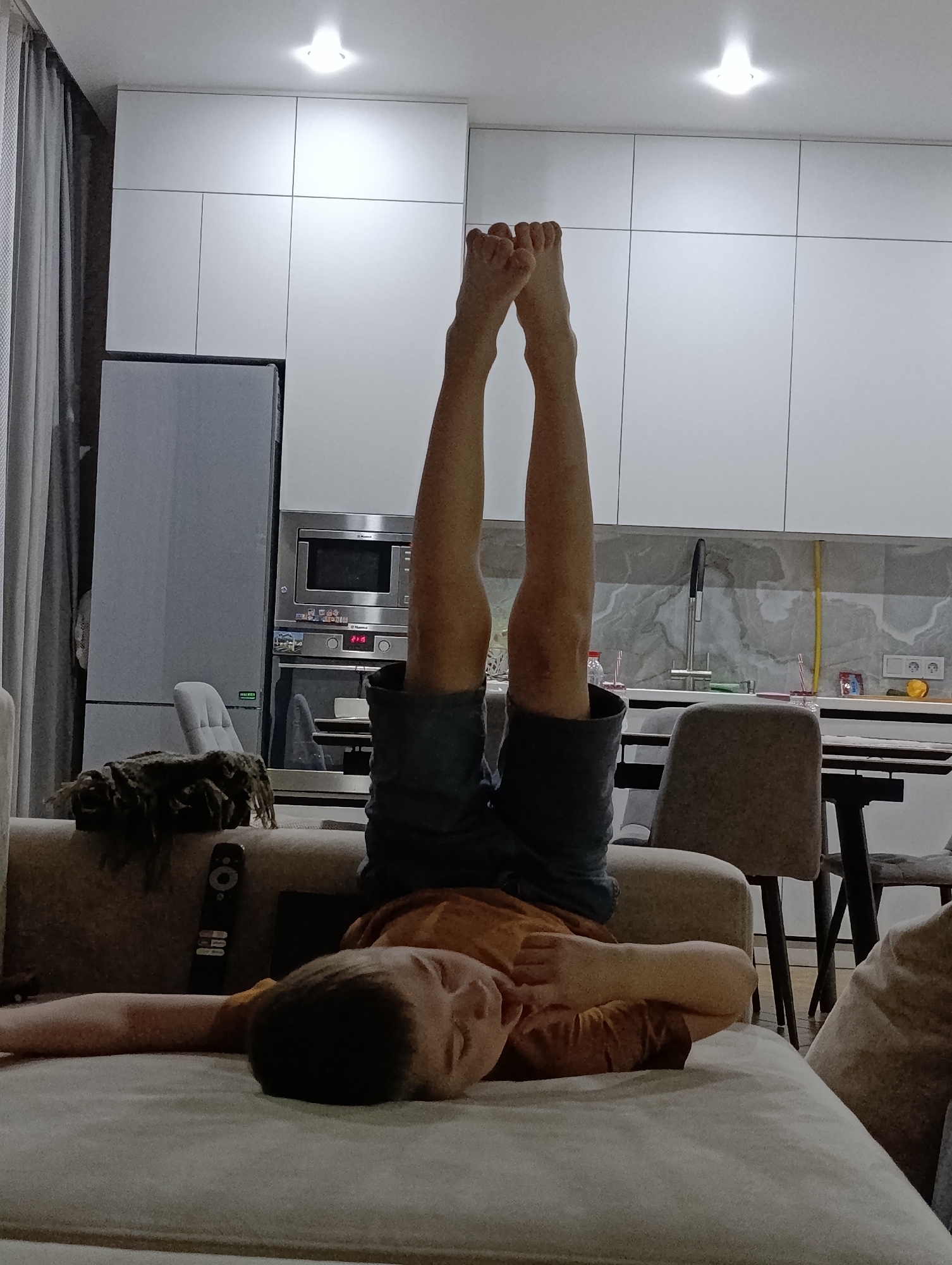 A boy lies on a beige couch in a modern-looking interior. His legs are raised vertically in the air, his feet visible. He wears a mustard-colored shirt and dark shorts. A remote control and a dark rectangular object rest on the couch near him. The background features a white kitchen with appliances, including a refrigerator, microwave, and sink. Gray curtains are to the left. The photograph was taken indoors, likely in a residential area in the city of Chaikovsky, Russia.