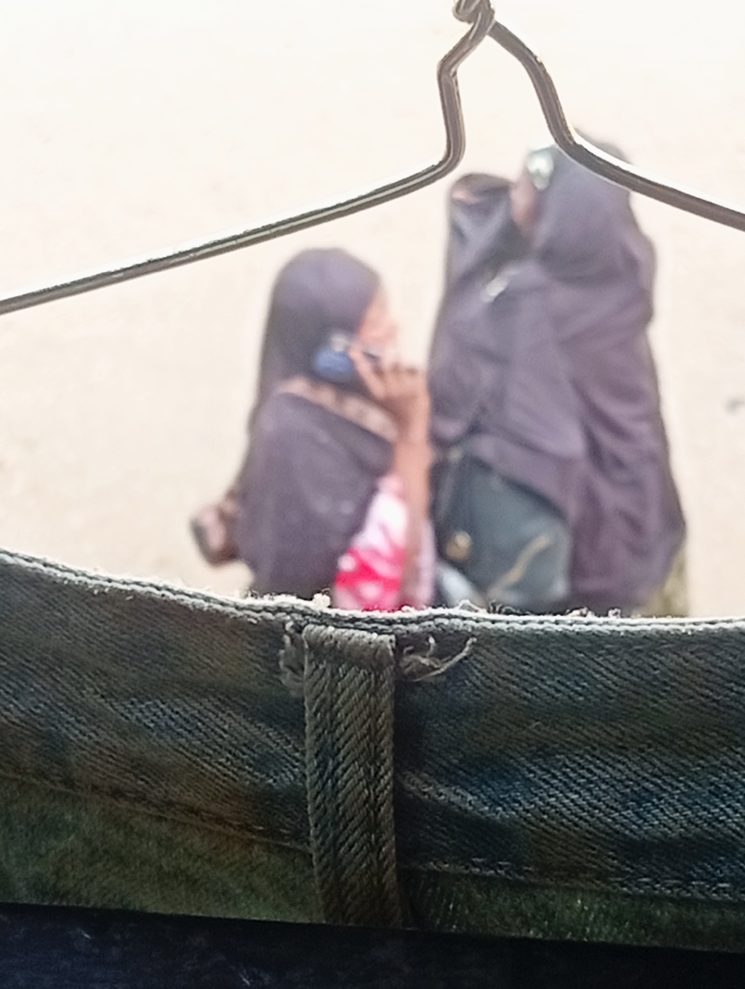 In Bukuru, Nigeria, two figures, one holding a phone to her ear, are seen through a metal hanger in the foreground. The subjects are out of focus, wearing what appears to be traditional clothing. The immediate foreground is dominated by the textured denim of clothing hanging on the hanger, with frayed edges suggesting wear. The background is a blurred, light-colored surface, possibly a wall or sky.