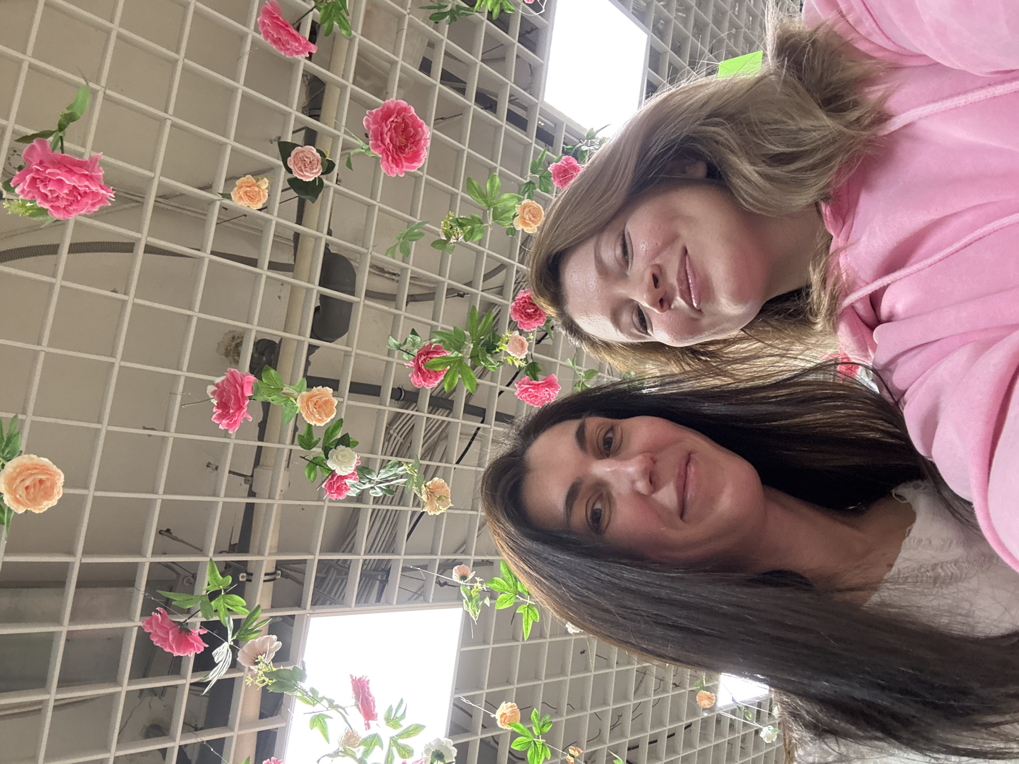 The image captures a vibrant indoor scene featuring two women smiling, positioned from a low-angle perspective. The woman in the foreground has long, dark brown hair and is wearing a light-colored top, while the woman behind her, slightly to the left, has lighter brown hair and is dressed in a pink hoodie. Both women are looking towards the camera with gentle smiles, suggesting a positive and relaxed mood, likely posing for a selfie or a casual photograph.
The background is dominated by a decorative white grid-like ceiling structure, possibly a trellis or an open-grid ceiling, from which numerous artificial flowers are suspended. These flowers are predominantly in shades of pink and peach, with some white accents, accompanied by green leaves, creating a festive or aesthetic environment. Bright square-shaped lights are visible within the ceiling grid, illuminating the scene and indicating an indoor location, possibly a retail space, a cafe, or an event venue in Votkinsk, Russia. The image is rotated counter-clockwise, giving it an unconventional, dynamic composition. There is no visible text in the image.