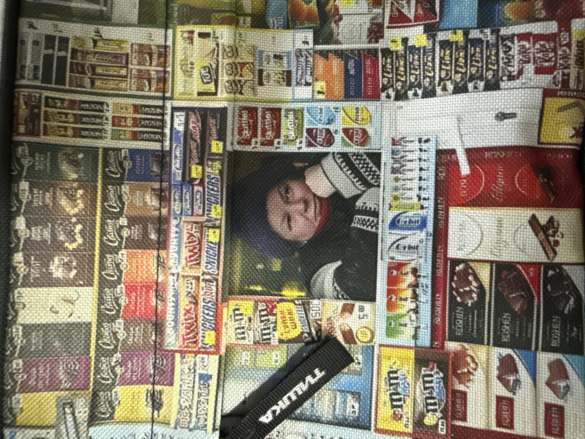 The image displays a close-up, overhead shot of a fabric surface covered with a repeating pattern of candy bar wrappers. The wrappers are colorful and feature various brand names and imagery associated with confectionery. Among the visible brands are "Twix," "Snickers," "M&M's," "Skittles," and "Roshen." The pattern is dense, with wrappers arranged in a grid-like fashion, creating a mosaic effect. The fabric appears to be a durable material, possibly from a bag or a similar item. No people, animals, or specific environmental features indicative of Novorossiysk, Russia, are present in the image. The scene is entirely composed of the printed candy bar wrapper pattern on the fabric.