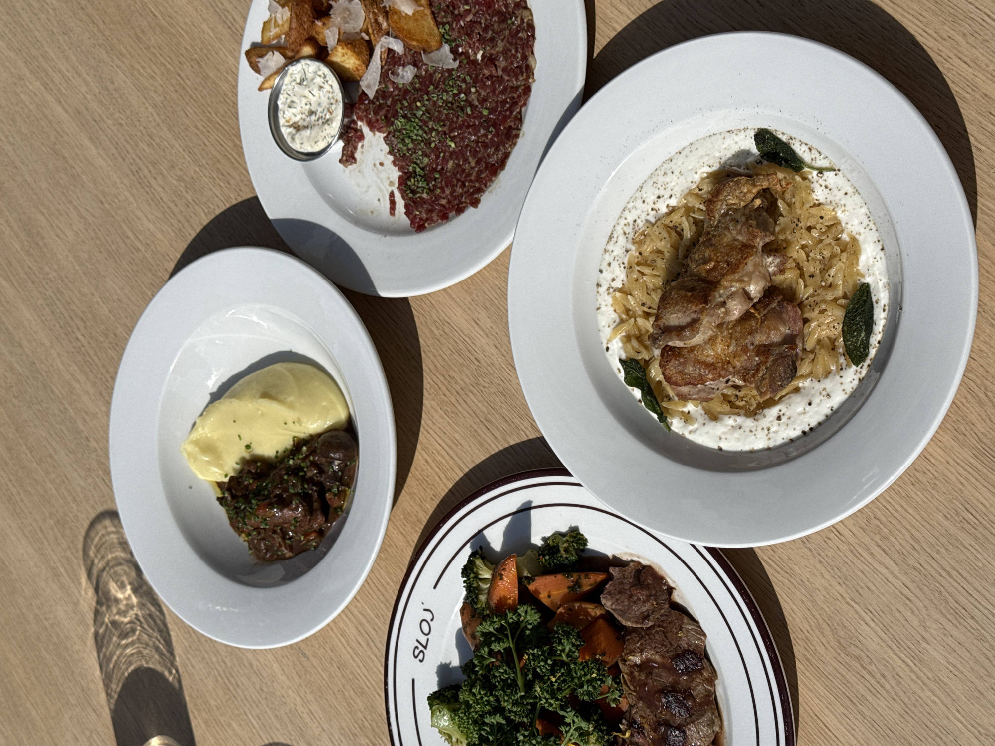 The image captures a top-down view of four distinct dishes served on a light wooden table, likely during a daytime meal at a restaurant.
On the top left, a white plate holds a dish of minced raw meat (possibly steak tartare) garnished with herbs, accompanied by crispy potato wedges or chips and a small metal cup of white dipping sauce, possibly aioli. Some grated cheese is also scattered over the potatoes.
The top right plate features a portion of braised or roasted meat, likely chicken or pork, resting on a bed of orzo pasta or rice pilaf, all covered in a creamy, speckled sauce and garnished with a few green sage leaves.
At the bottom left, another white plate presents a hearty, dark stew, possibly beef or lamb, next to a generous serving of smooth yellow mashed potatoes or polenta, topped with finely chopped green herbs.
The bottom right plate, which has a distinctive brown and white striped rim, displays slices of grilled meat (possibly steak or lamb) alongside roasted vegetables including carrots and broccoli florets, and a fresh sprig of parsley. The word "SLOJ" is visible in an arc along the plate's rim.
Strong, distinct shadows are cast across the table and plates, indicating bright natural sunlight and suggesting the time of day is mid-morning or afternoon. The overall scene depicts a restaurant dining experience in Belgrade, Serbia.