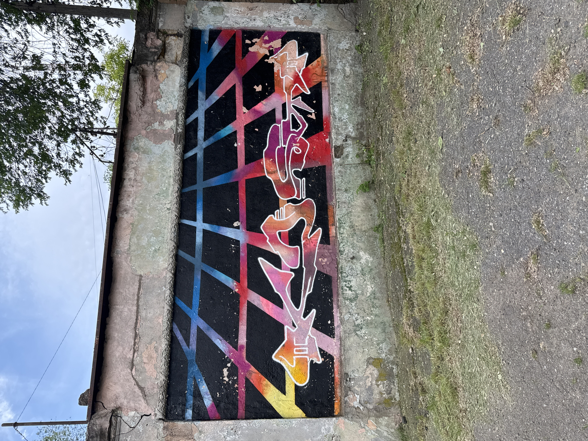 This image, taken in Odesa, Ukraine, depicts a weathered exterior wall featuring a large graffiti mural, with ground cover in the foreground and a cloudy sky visible above.
The central subject is a rectangular graffiti piece painted on a deteriorated wall structure. The graffiti's background consists of a black base over which a perspective grid of diagonal lines is painted. These lines exhibit a gradient color scheme, transitioning from blue and purple in the upper left, through pink and red in the center, to orange and yellow in the lower left and right. Overlaid on this grid are stylized, angular letters or shapes with white outlines, filled with a multi-color gradient (orange, pink, purple, red, brown). The paint on the black background shows areas of peeling and flaking, particularly on the right side, revealing the underlying wall surface.
The wall surrounding the graffiti is constructed of concrete or stucco over masonry, exhibiting significant signs of age and neglect. Its surface is extensively cracked, with large sections of paint and plaster peeling away, exposing varied textures and colors (pale pink, light green, and grey). A prominent vertical crack runs down the upper left section of the wall. Along the top of the wall, a rusted horizontal metal beam is visible. Above this beam, the wall cap is uneven and appears to be concrete or stone. On the right side, exposed rebar or wires protrude from the wall's top edge, and strands of barbed wire are present.
Above the wall, a partially overcast sky is visible. To the left, a slender metal post or utility pole extends upwards, with a single wire stretching across the sky. On the right, a deciduous tree with green foliage partially obscures additional utility poles and wires.
The ground in the immediate foreground is composed of a patchy mixture of asphalt or dark, coarse pavement and sparse, short grass and weeds. Small bits of dried plant matter and debris are scattered across the ground surface. The pavement shows signs of wear and minor irregularities. No people are present in the scene, and no overt actions or interactions are depicted.