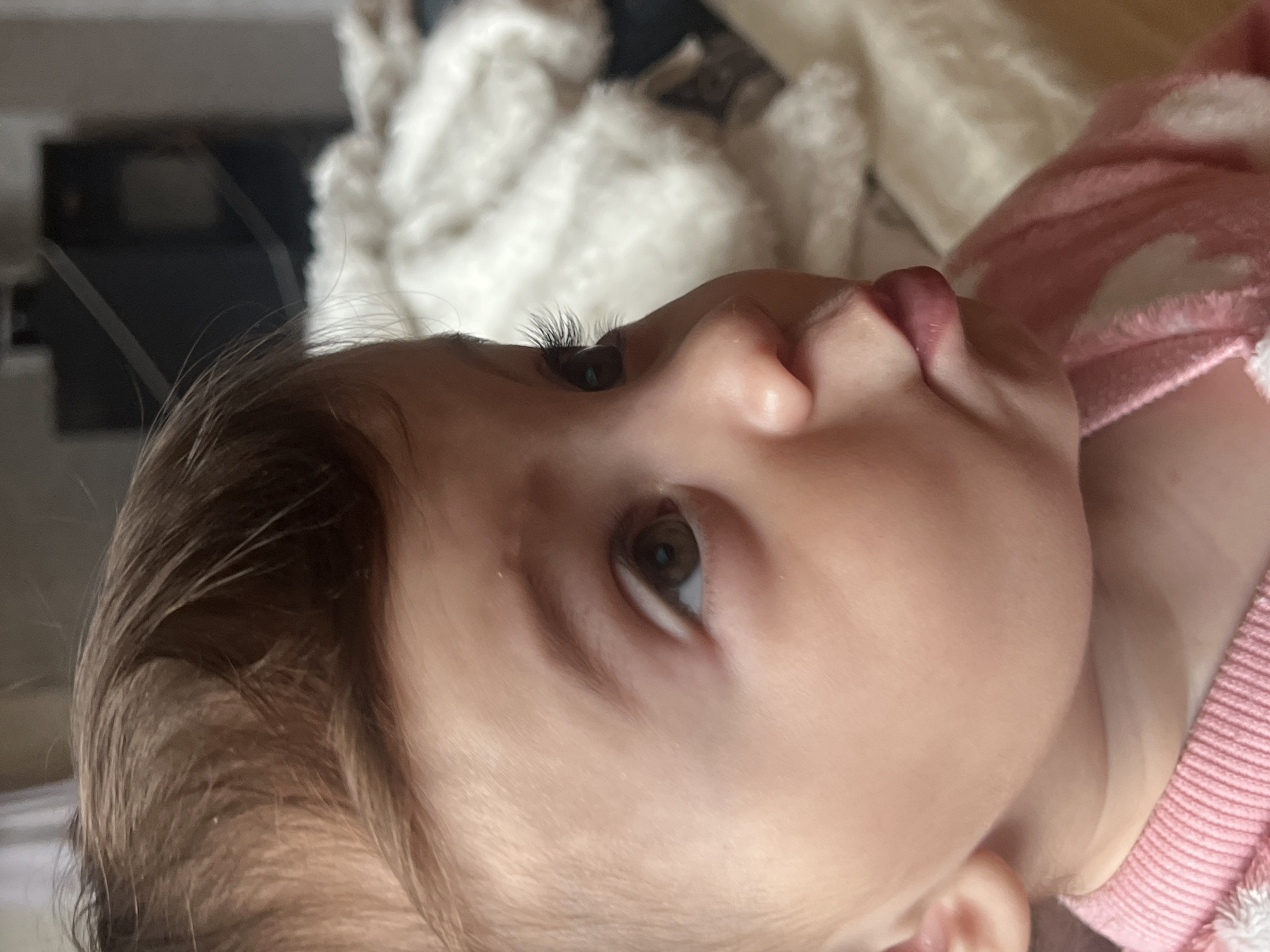 A close-up, eye-level shot shows a baby or toddler lying down, looking upwards. The child has fair skin, light brown hair, and hazel eyes. The child's mouth is slightly open. The child wears a pink and white patterned shirt. In the background are a white, fluffy blanket and a blurred dark object. The location of Elena, Bulgaria is not discernible from this image.
