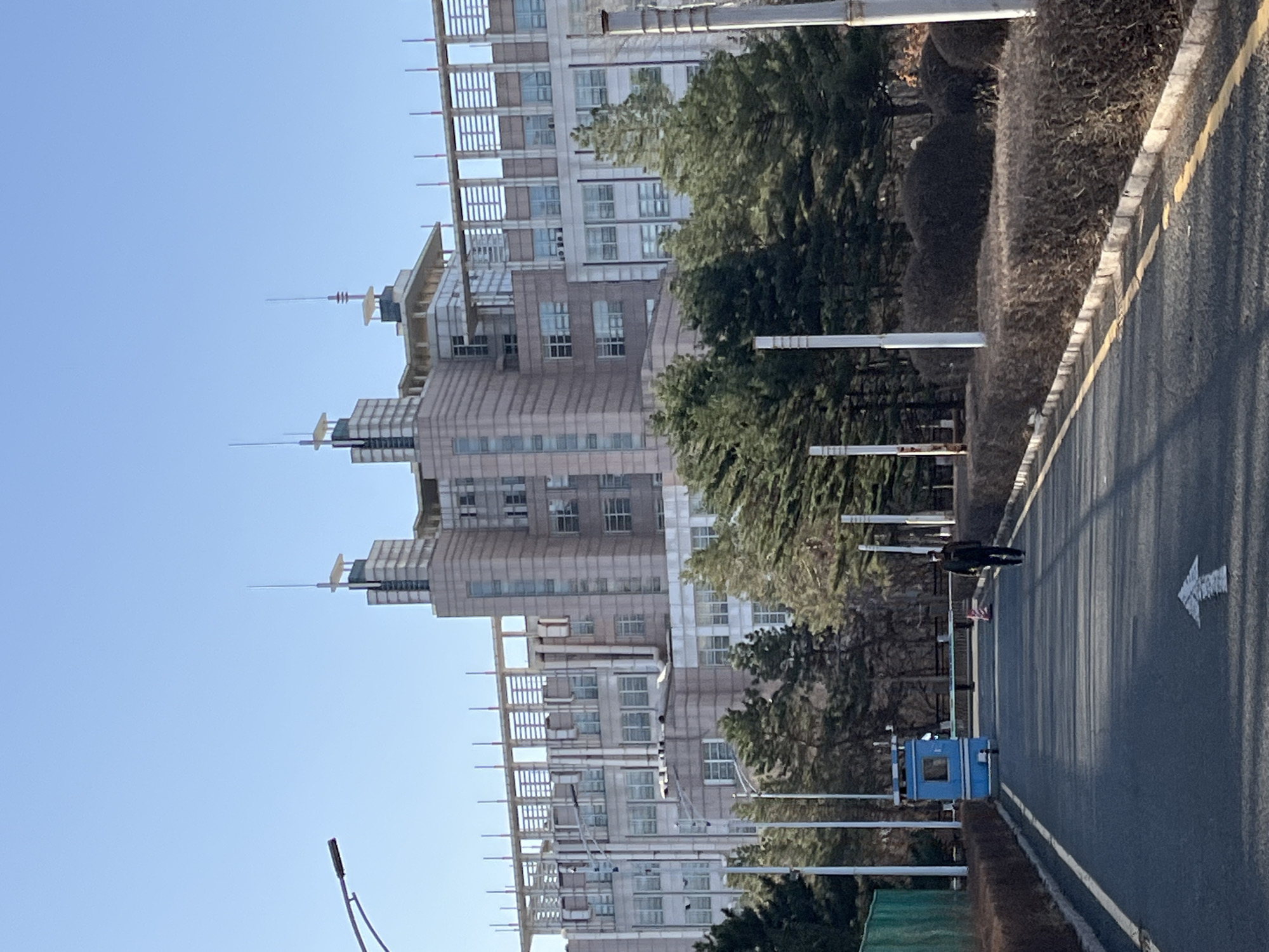 The image shows a scene in Changchun City, China, featuring a modern, multi-story building with a distinct architectural style. The building is light-colored with numerous windows and decorative elements on its roof. The building is flanked by green trees and shrubs. A paved road runs parallel to the building and is marked with a white arrow indicating direction. A blue utility or control box sits along the side of the road, and a figure is walking along the roadside. The sky is clear and blue.