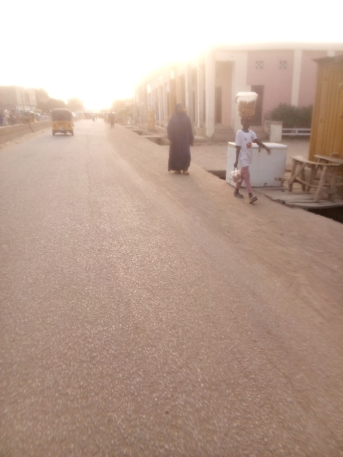 The image depicts a sun-drenched street scene in what appears to be Tudun Murtala, Nigeria, likely during the early morning or late afternoon given the bright, somewhat hazy light and the long shadows. The sun, positioned towards the horizon, casts a strong glare, washing out parts of the sky and illuminating the scene.
The foreground features a wide, paved road, appearing slightly sandy or dusty, with a coarser, unpaved shoulder or dirt path running alongside it to the right.
Several individuals populate the scene. On the right side of the road, closer to the viewer, a person dressed in a long, dark blue or black garment stands, facing away from the camera. Further along the right, a younger person, possibly a child or young adult, walks away from the camera. They are wearing a light-colored t-shirt and pink trousers, skillfully balancing a large, clear container filled with what appear to be food items (perhaps fried snacks or bread) on their head, and carrying a white bag or package in their left hand. This suggests they might be a street vendor or transporting goods.
In the mid-ground, on the left side of the road, a yellow auto-rickshaw, commonly known as a Keke Napep, is visible, driving away from the viewer. Other indistinct figures can be seen further down the road, indicating a degree of activity.
The right side of the street is lined with buildings, some with simple facades and windows or doorways. Closer to the right edge of the frame, there's a white, rectangular object (possibly a cooler or a small stall base) and several wooden structures, which could be tables or makeshift market stalls, further emphasizing a commercial or community setting.
The overall atmosphere conveys a snapshot of daily life and routine activity under a bright, clear sky. No discernible text is visible on any signs, clothing, or objects in the image.