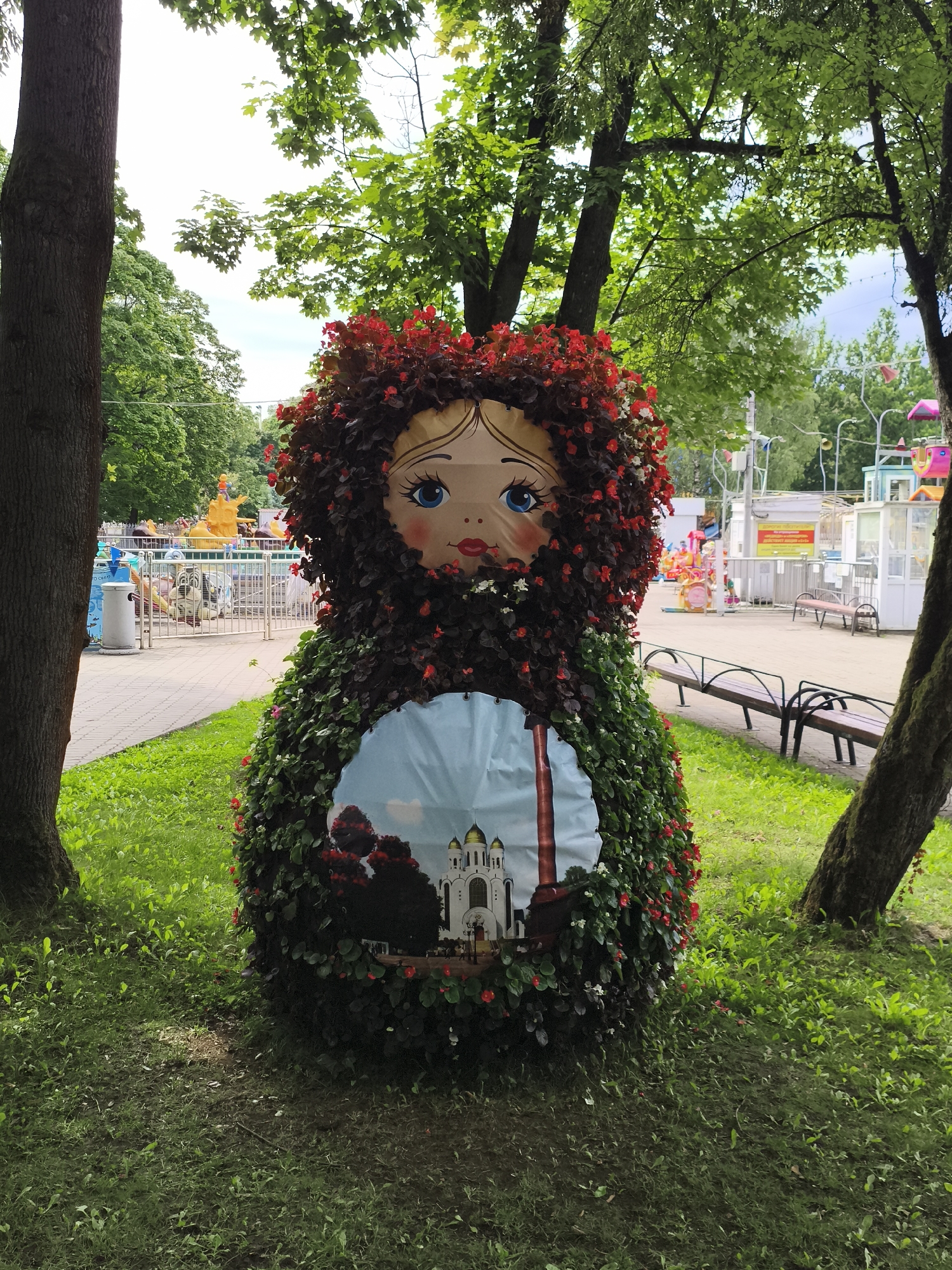 A large, doll-shaped topiary resembling a matryoshka doll is situated in a grassy area in front of a park or amusement area. The doll is adorned with numerous red and white flowers, forming its "hair" and the body. The face of the doll, with blue eyes and red lips, is painted on a flat surface protruding from the floral structure. A circular panel on the lower half of the doll's body depicts a stylized image of a white cathedral with golden domes, possibly a landmark in Kaliningrad, Russia, given the context. In the background, the park features children's rides, benches, pathways, and trees. The overall scene suggests a public outdoor space designed for recreation and possibly decorated for a festival or seasonal event.
