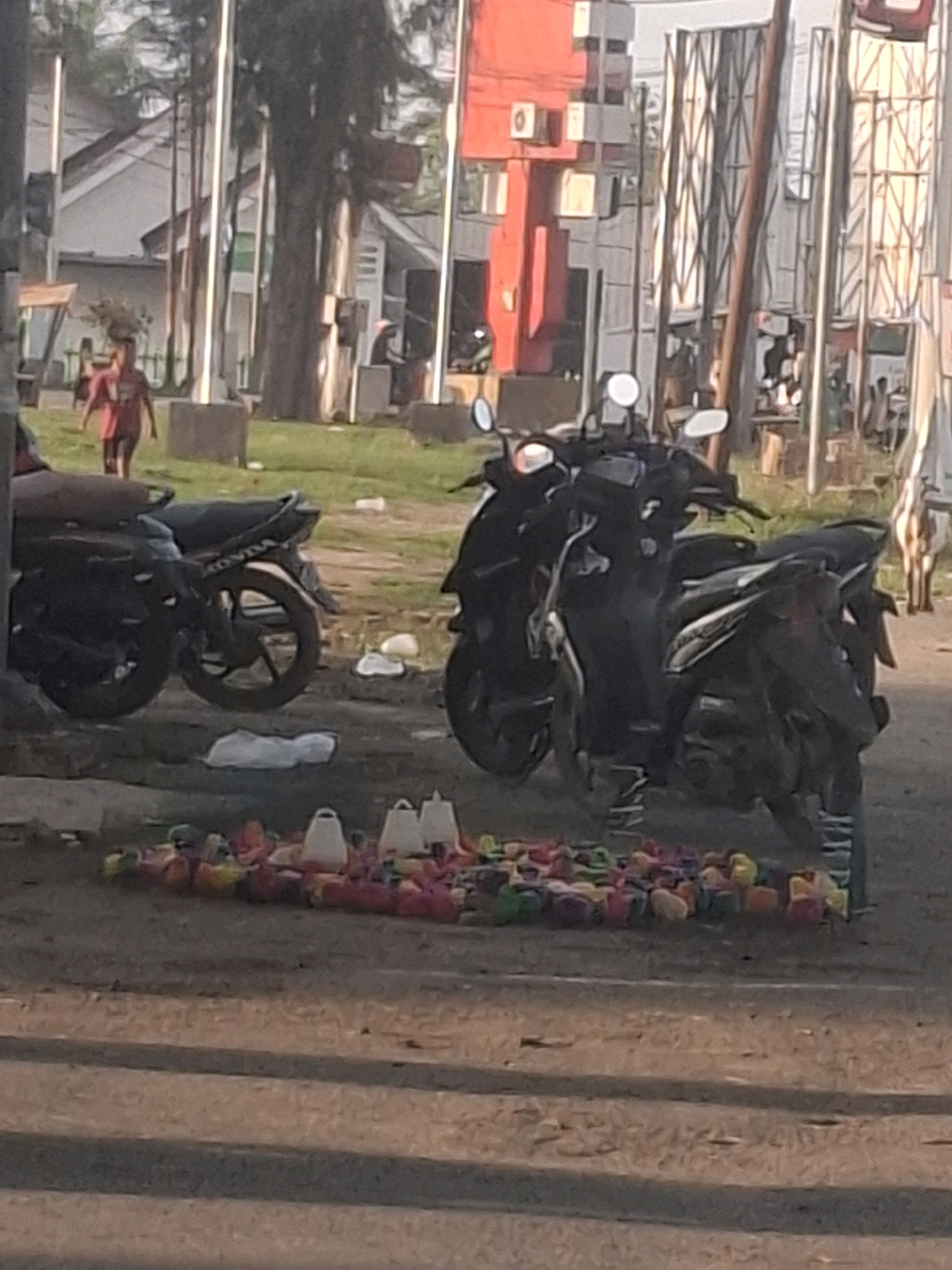 The image shows a street scene in Mesjid Tuha, Indonesia, during daylight. In the foreground, there is a collection of colorful plastic toys and two white objects, possibly feeders, arranged on the ground. Behind these items, two dark-colored motorcycles are parked. The motorcycle on the left is partially obscured by the one on the right, which is more fully visible and appears to be a scooter. Further back, a young person wearing a reddish shirt is walking away from the viewer towards a tree-lined area. In the background, buildings with red and white facades are visible, along with metal fencing and utility poles. The environment appears to be a mixed urban and semi-rural setting.