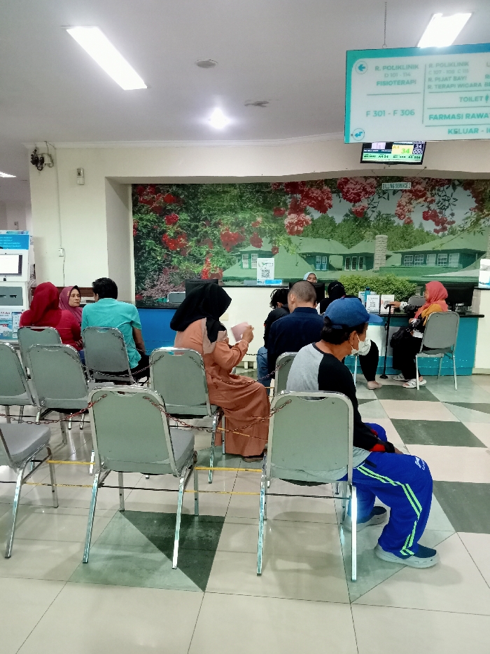 The scene is a waiting area in a hospital, located at coordinates (-6.76, 111.015) in Kecamatan Margorejo, Indonesia. Seven individuals are seated in rows of chairs, facing a reception desk. The room features a tiled floor with a checkered pattern. The wall behind the chairs displays a mural of a lush green garden with red flowers, a white picket fence, and a small white house. A sign above the reception desk indicates that it is a clinic offering physiotherapy services. There is a digital clock displaying the time 09:43:02. A blue sign with white text announces that there are a variety of services offered in the building, including physiotherapy, a pharmacy, and a toilet. There is a metal railing separating the waiting area from the reception desk. The waiting area is brightly lit with fluorescent lights. Some of the individuals seated are wearing face masks, suggesting the scene could be during a pandemic or a period of time when masks were required in public spaces. The subjects in the photo are all of Asian ethnicity. There are no discernible interactions happening between individuals. The scene appears to be an everyday occurrence in the hospital.