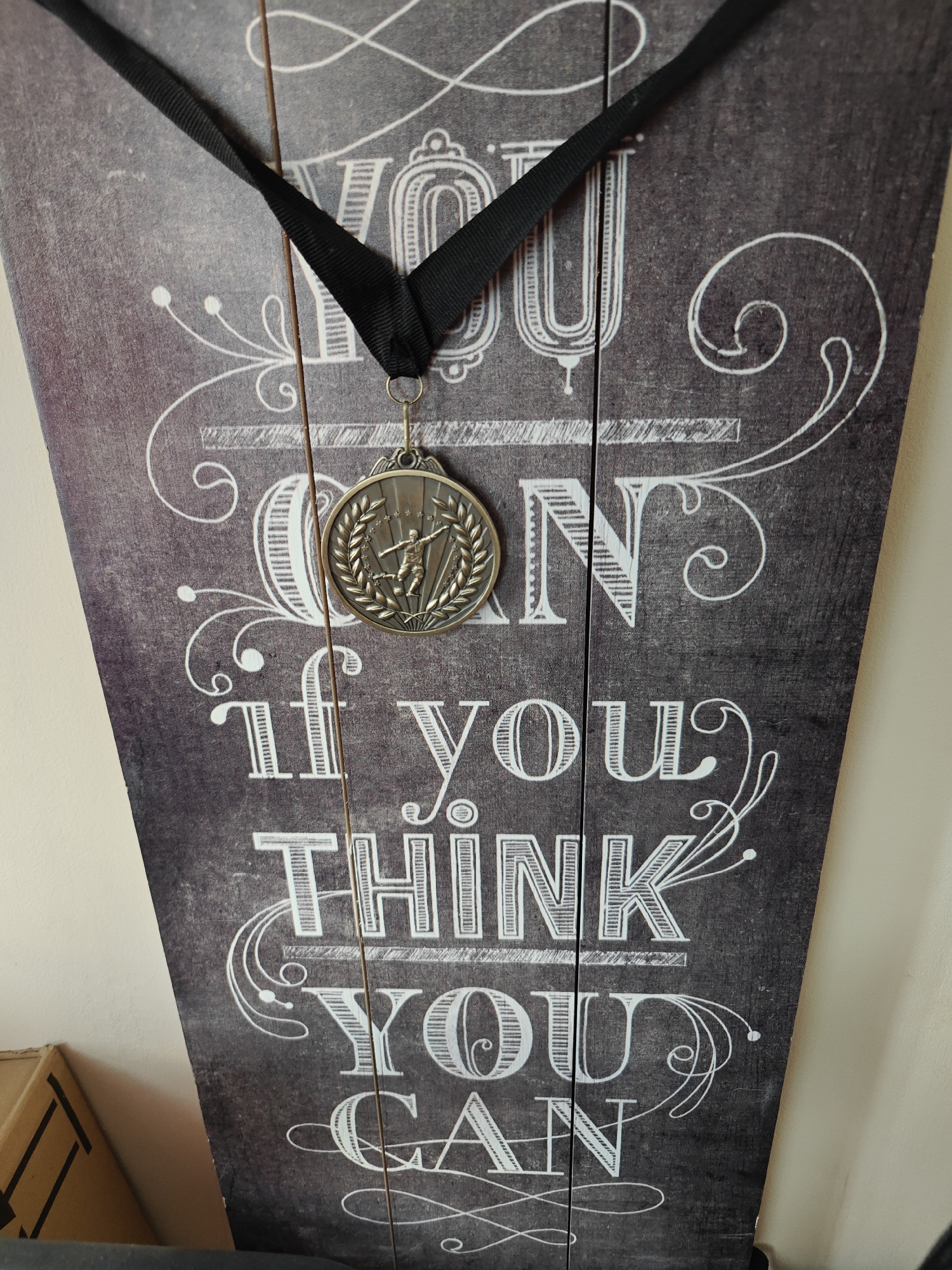 The image shows a close-up view of a medal hanging on a decorative wooden sign. The sign is dark gray with white lettering that reads "YOU CAN IF YOU THINK YOU CAN" in stylized, decorative font. A dark, likely black, ribbon suspends the medal, which is circular and bronze-colored. The medal features an embossed design depicting a figure in a dynamic pose, possibly related to sports or athletics, surrounded by a laurel wreath. The wooden sign appears to be constructed of multiple planks, visible by the vertical lines running along its surface. The scene is indoors. There are no explicit indicators that the scene is in Maidstone, United Kingdom.