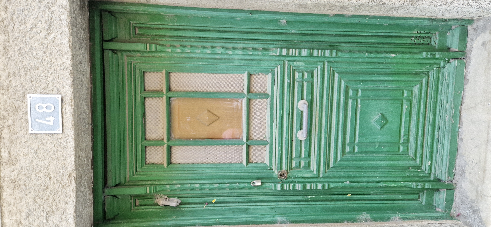 A green wooden door with a metal handle and a padlock is visible. Above the door, a house number "48" is displayed on a gray plaque attached to a textured stone wall. The door has intricate carvings and a glass panel with a diamond-shaped wooden insert. Some white residue is present on the door and wall.