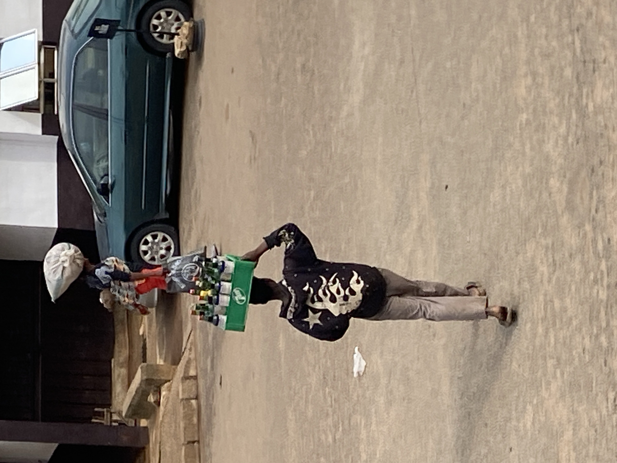 The image shows a person standing on a paved surface, possibly a street or sidewalk, in Bukuru, Nigeria. The person is carrying a green crate filled with bottles, which appear to be beverages. They are wearing a dark-colored shirt with a flame-like design and stars, and light-colored pants. On their head, they are carrying a large, light-colored, possibly cloth-wrapped item, balanced precariously. To the left of the person is a dark green car. The setting appears to be outdoors, with a building structure to the left, and the ground is a rough, textured pavement. The lighting suggests it is daytime, with some shadows visible. The overall scene depicts a person engaged in street vending or transportation of goods. No visible text is present in the image.