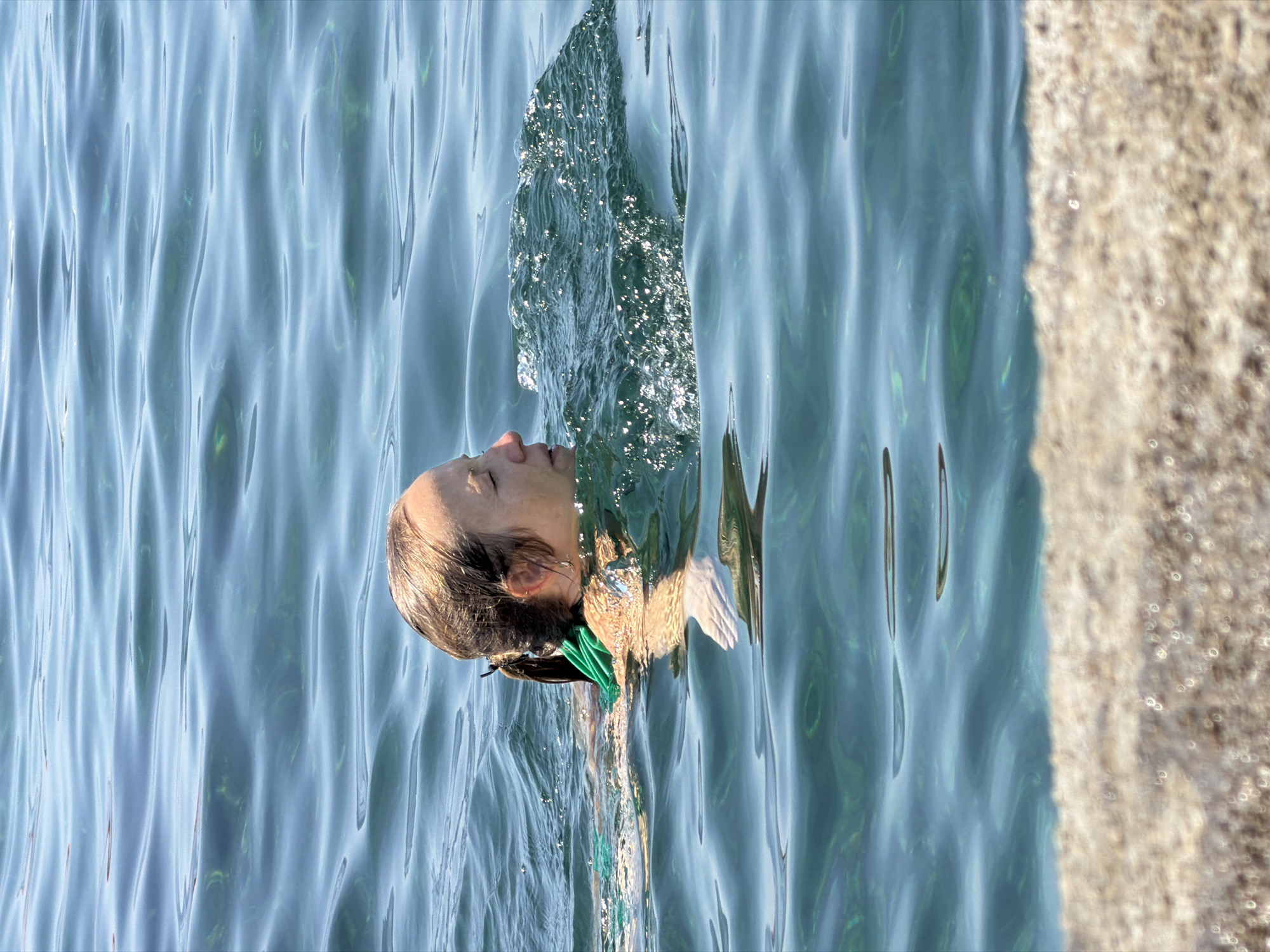 A medium shot captures a woman swimming in clear, turquoise water. Her head is partially submerged, with her face turned to the right, eyes closed. Her brown hair is pulled back in a ponytail and appears wet. She is wearing a green swimsuit. The water exhibits gentle ripples and sparkles in the sunlight, indicating a bright day. In the foreground, a blurry, light brown surface, possibly a rock or the seabed, is visible. The overall impression is of a serene moment during a swim in a coastal environment, likely the Adriatic Sea near Budva, Montenegro, given the characteristic clarity and color of the water.