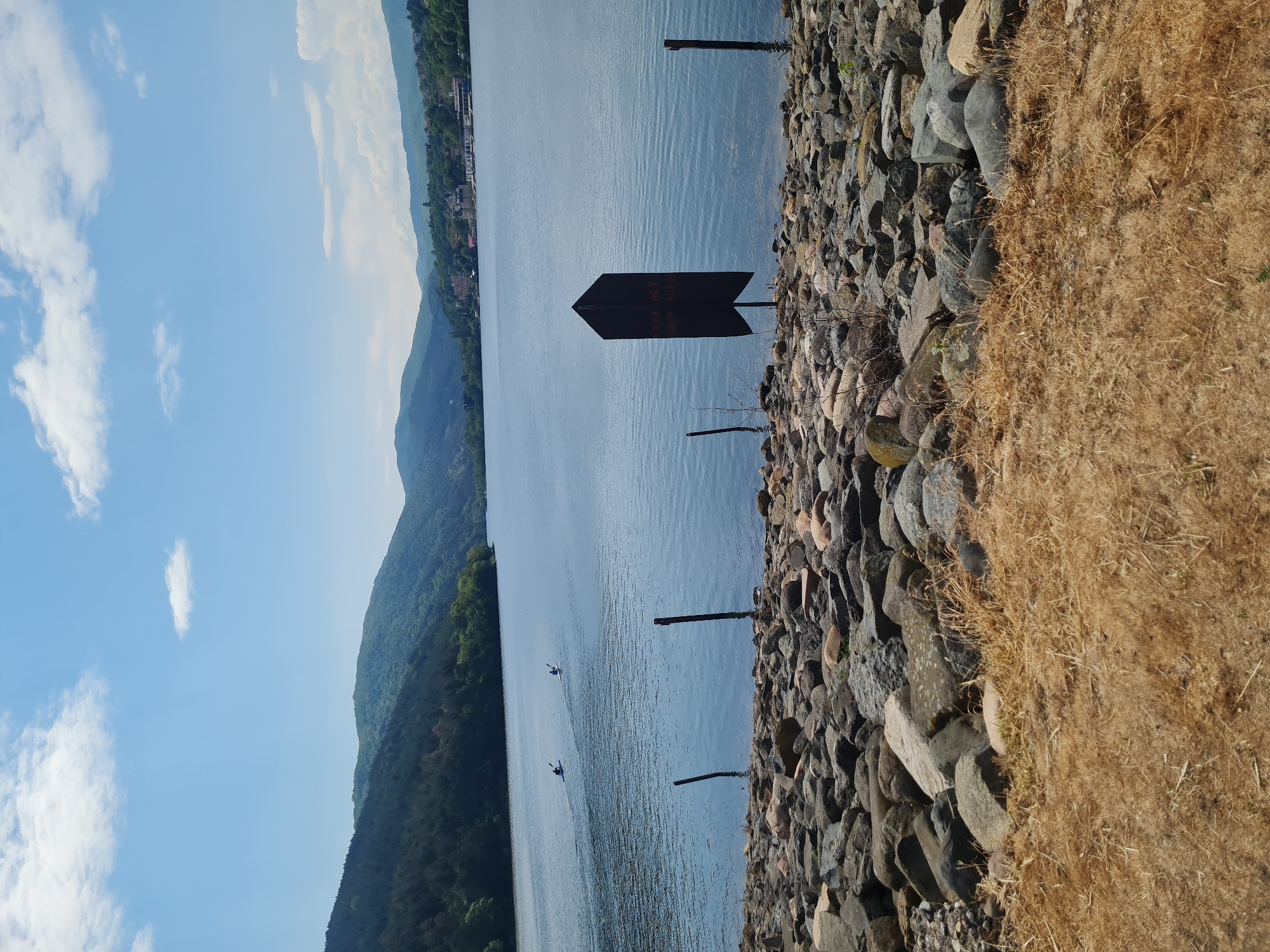 The image depicts a view of Lake Pancharevo in Pancharevo, Bulgaria. A body of water fills the majority of the frame, flanked by a rocky shoreline on the right and mountainous terrain on the left. Two jet skis are visible on the lake's surface. Several wooden posts stand along the shoreline, with a dark, arrow-shaped sign fixed to one of them. The foreground to the right of the frame features dry, yellowish grass. The sky is partially clear with scattered white clouds. The mountains in the background are covered in greenery and residential or commercial structures are visible further up the mountains.