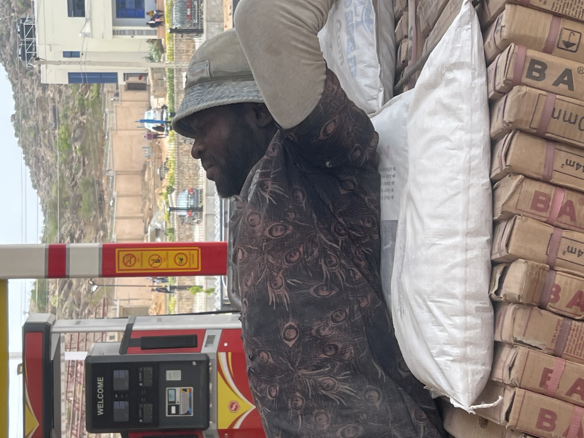 The image depicts a man carrying goods in an outdoor setting, likely a commercial or roadside area, in Jos, Nigeria. The man, dark-skinned with a beard, wears a light-colored bucket hat and a dark shirt patterned with what resembles peacock feathers. He is positioned in the foreground, facing right, with his left arm raised, seemingly supporting a large white sack on his back or shoulder.
To his right, a stack of cardboard boxes is visible, bound together with reddish straps. These boxes have "BA" and "4m³" (or possibly "4mm") printed on them. Behind him and to his left, a red and white barrier is present, adorned with yellow warning signs that are difficult to read precisely. Below the barrier, a red and yellow fuel pump or similar machine is visible, displaying "WELCOME" text and a digital interface.
The background shows a light-colored building with blue window frames. Beyond this structure, a rugged, rocky hill with sparse vegetation rises, suggesting a dry or semi-arid environment. The lighting indicates daylight, possibly bright but slightly overcast, as shadows are not distinct. The man appears focused on his task, with no discernible emotional expression. The scene suggests a busy or working environment, with goods being moved or delivered.