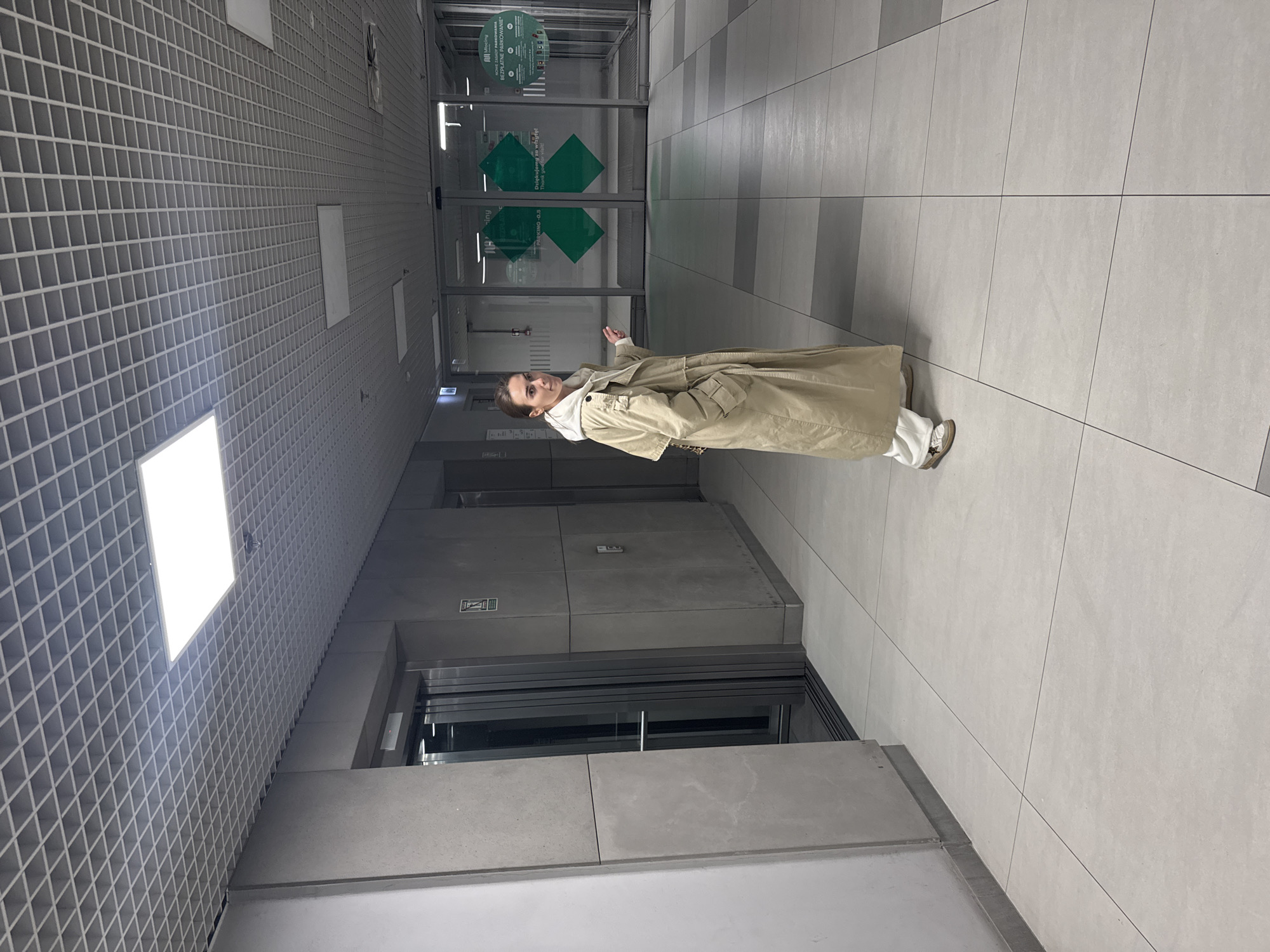 An indoor, modern hallway or corridor is captured in a portrait-oriented shot, featuring a person standing centrally. The subject, a woman, wears a light beige trench coat over a white hoodie and white pants, paired with light-colored sneakers. She is looking towards the camera with a slight smile, her right arm gently raised.
The setting appears to be a public building, possibly a transportation hub or a shopping center, given the "P+R" (Park and Ride) and "Młociny" signage. The floor is covered with large light-grey tiles, some featuring darker grey stripe patterns. The ceiling is a distinctive grid of light grey squares, illuminated by several large, bright, square fluorescent light panels. To the person's left are two elevator doors with metallic finishes.
In the background, behind the person, are automatic glass sliding doors with green diamond-shaped signs. Visible text on these signs includes "Mlociny" and "P+R," indicating a location in Warsaw, Poland. Other text in Polish and English reads: "Dziękujemy za wizytę! Thank you for visit," and mentions "BEZPŁATNE PARKOWANIE" (free parking). The overall impression is a clean, well-lit, contemporary space. The person is actively posing for the photograph.