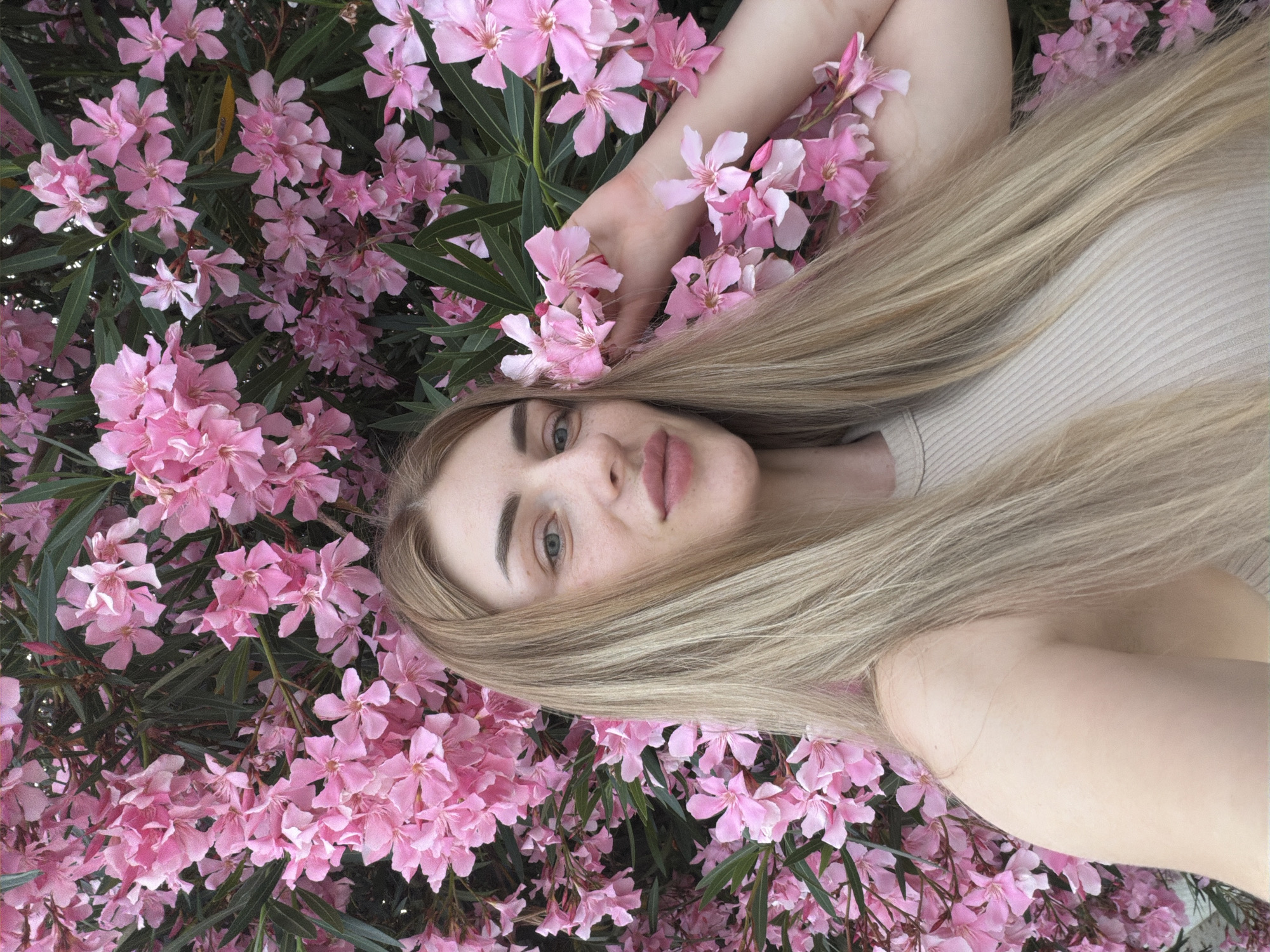 In an outdoor setting in an unknown city, a young woman with long, blonde hair stands in front of a dense bush of vibrant pink oleander flowers. She is facing the camera and wearing a light-colored, ribbed tank top. Her left arm is raised, with her hand gently touching some of the flowers near her face. The background is filled with the lush foliage and numerous blossoms of the oleander bush, creating a colorful and slightly obscured environment. The lighting suggests it is daytime.