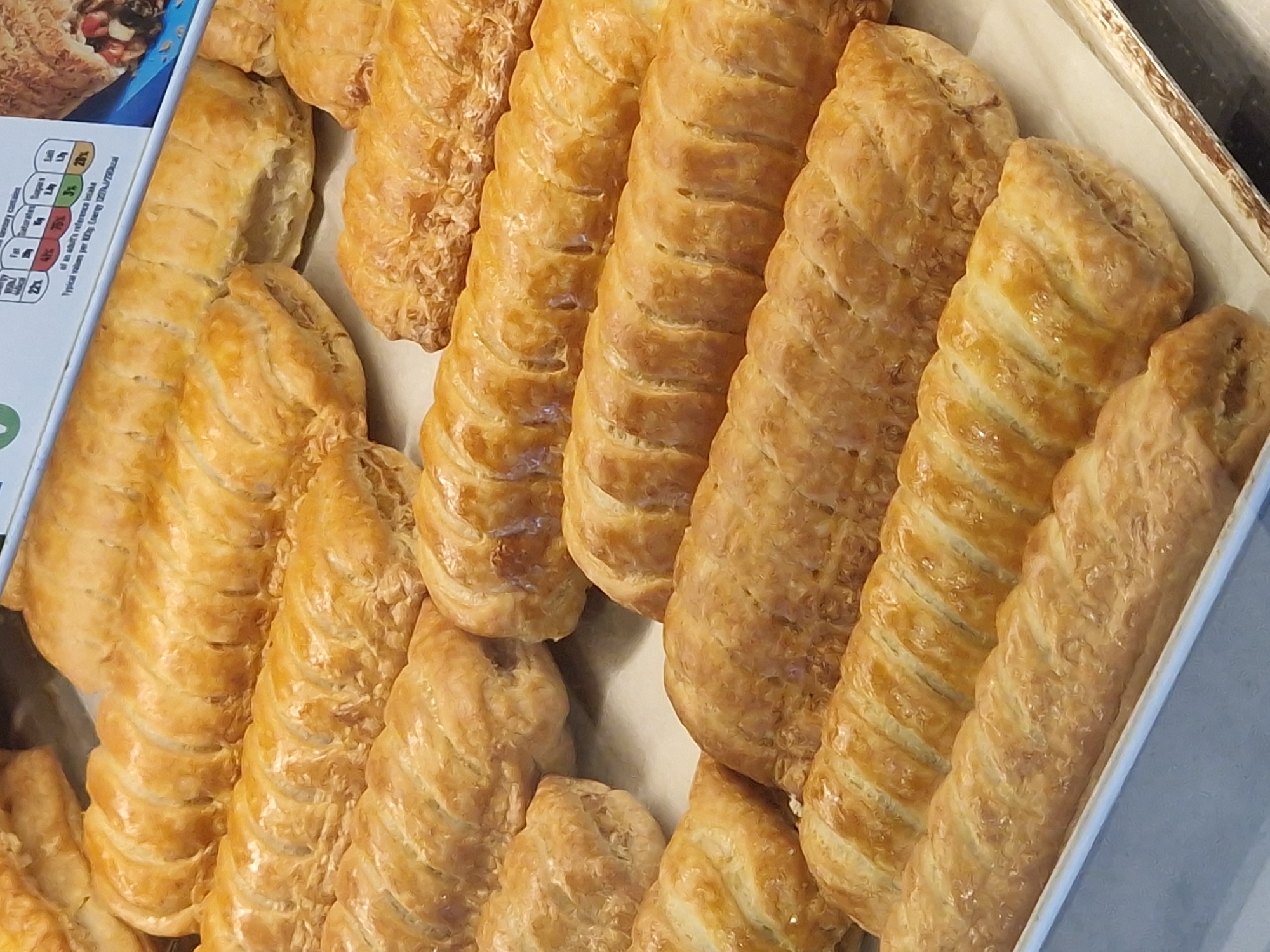 The image shows a tray of freshly baked sausage rolls. They appear to be arranged in neat rows within the tray, their golden-brown crusts and visible pastry layers suggesting a flaky texture. There are several sausage rolls in varying lengths and thicknesses. The tray seems to be made of a light-colored material, possibly cardboard or aluminum. The top left portion of the image contains a partially visible blue and white box with nutritional information. The location is unspecified and there is nothing to indicate the city of Pity Me, United Kingdom.