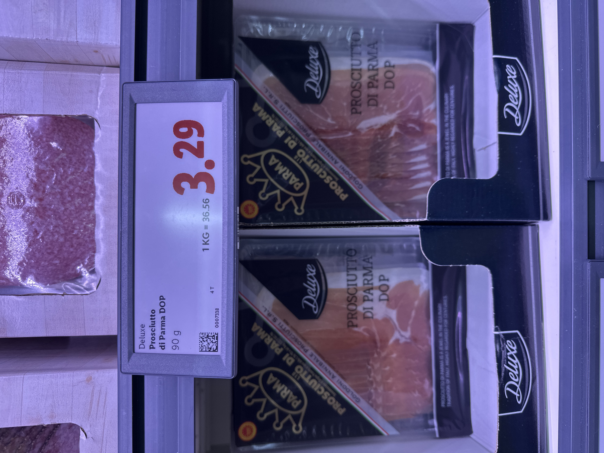 The image shows a close-up view of a refrigerated display case in a grocery store, likely in Bruges, Belgium. The focus is on two packages of "Prosciutto di Parma DOP" from the brand "Deluxe." The price displayed for the product is 3.29. The packages are in the foreground, with the electronic price tag visible above them. The price tag displays the product name, weight (90g), and unit price (1kg = 36.56). The background shows other products in similar packaging, suggesting a wider selection of deli meats. The lighting is artificial, typical of a supermarket environment. There are no people or animals visible in the frame.