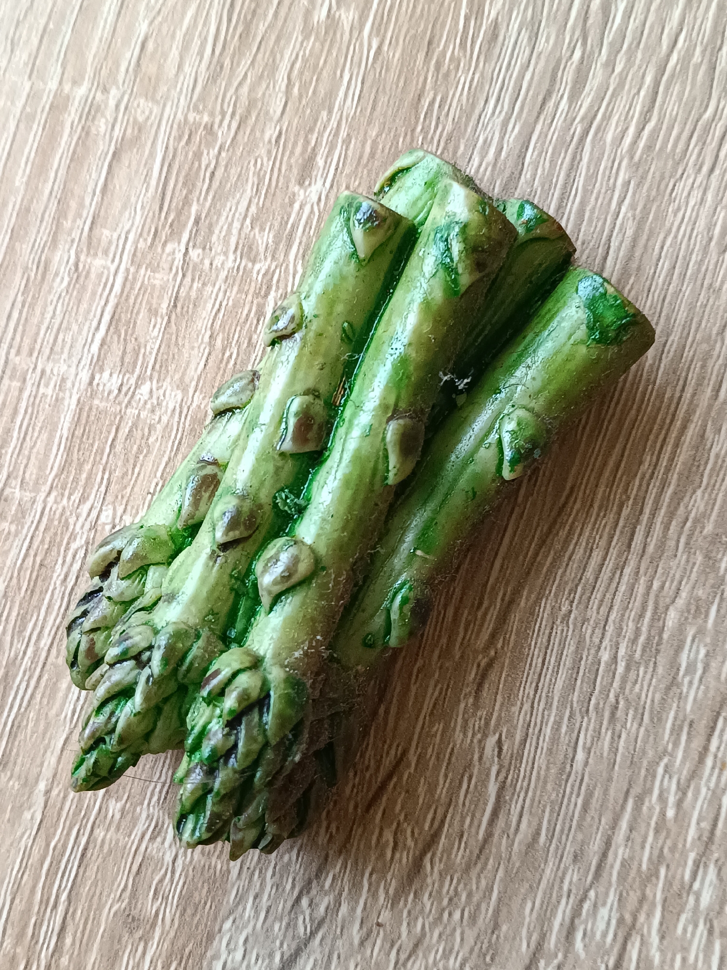 A cluster of five asparagus spears lies diagonally on a light brown wooden surface. The spears are green with small, brown, bud-like structures along their length. The wooden surface has a pronounced wood grain pattern running vertically from top to bottom. The image is a close-up shot, with the asparagus occupying the majority of the frame. There are no people or any other objects visible. The lighting appears to be from above and slightly to the left, casting faint shadows. The scene is located in Tebras, Latvia.