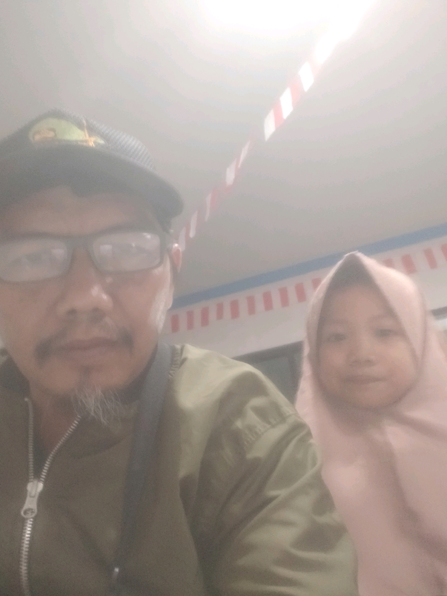 The image is a selfie taken by an adult male wearing glasses and a cap. He is positioned on the left side of the frame, while a young girl wearing a pink hijab is on the right. The background appears to be an indoor setting with a ceiling decorated with red and white stripes. The overall image is slightly overexposed, giving it a hazy appearance. No specific location within Sukaasih, Indonesia can be determined from the visual information provided.