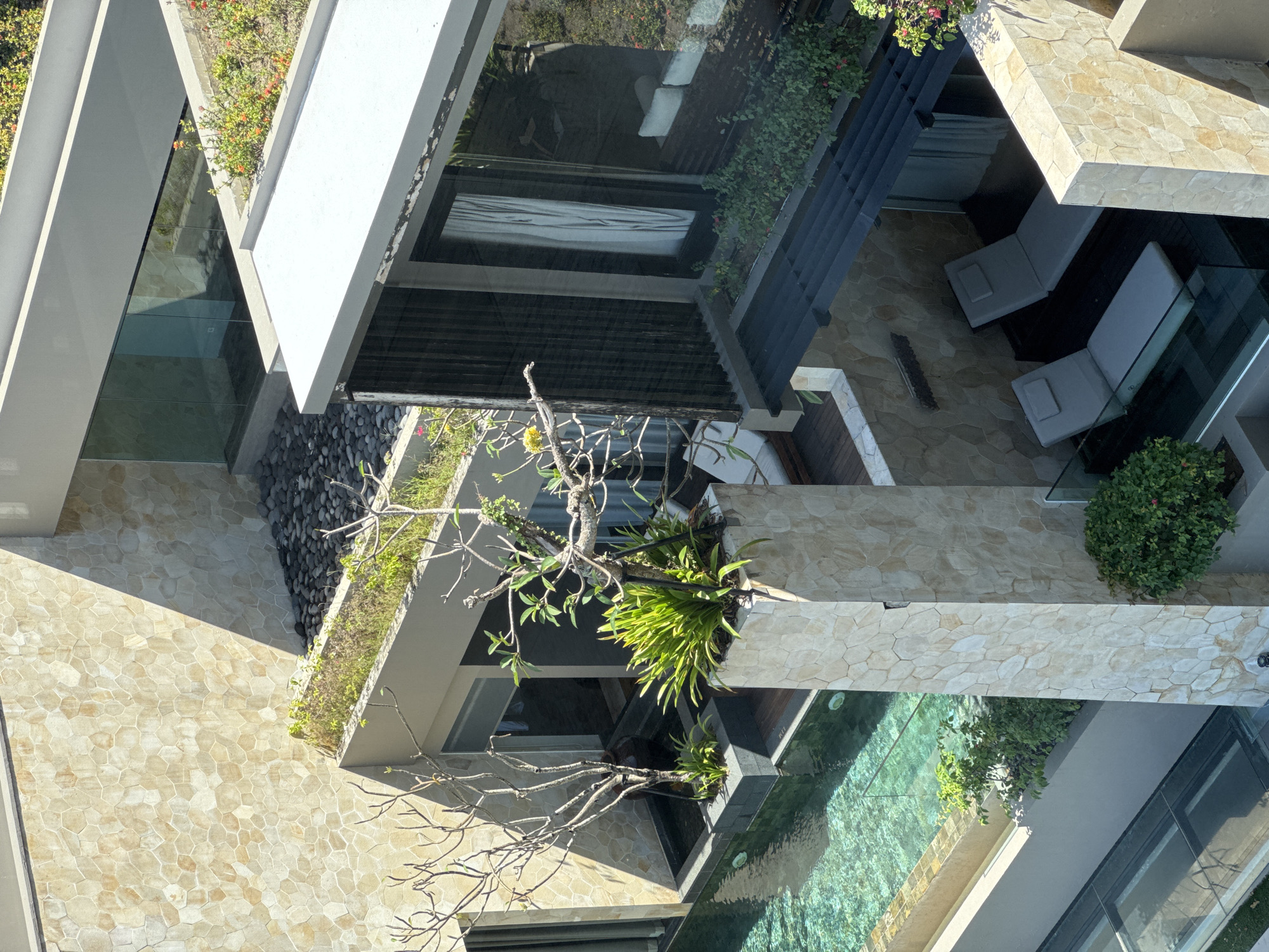 This aerial shot captures a contemporary multi-story building in Kuta Selatan, Indonesia, likely a resort or villa. The architectural design incorporates stone-tiled facades, concrete structures, and glass elements. Several levels feature green roofs or planters with various plant species, including mature trees with bare branches. A swimming pool with turquoise water is visible on one level. Below the pool, other parts of the building include lounge chairs and a bedroom with curtains. The lighting indicates it's daytime.