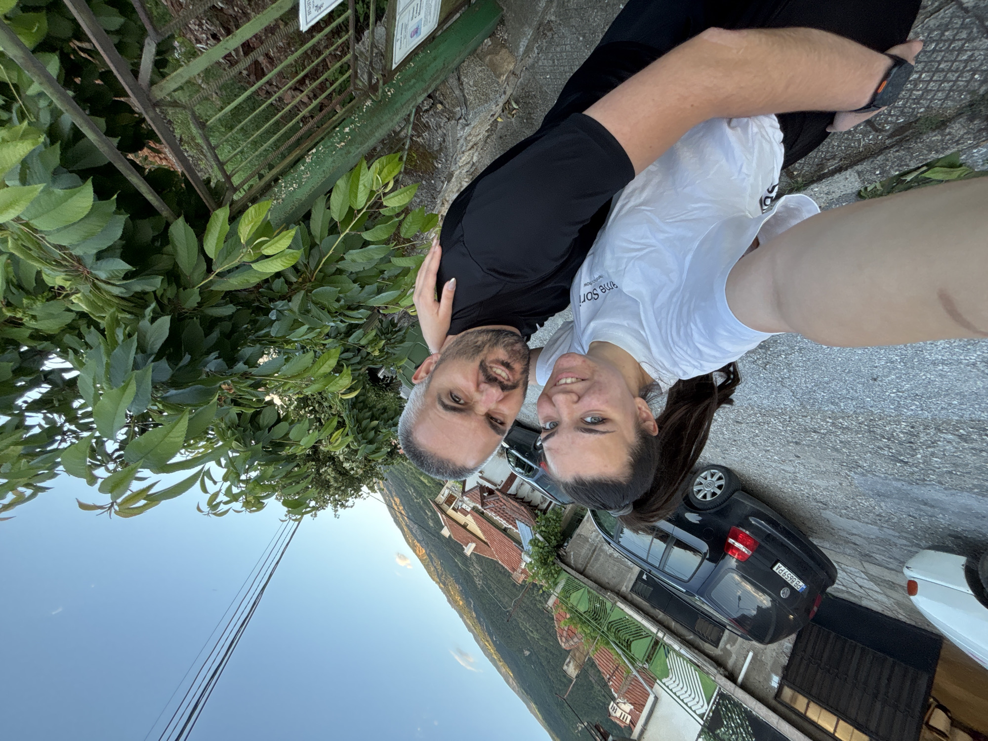 A selfie captures two individuals, a woman and a man, posing on a paved outdoor path in Karlovo, Bulgaria. The woman, with dark hair in a high ponytail and wearing a white t-shirt, smiles at the camera, holding her left arm extended for the shot. Her right arm is around the man. The man, with short grey hair and a beard, wearing a black t-shirt, makes a humorous grimace towards the camera with his right arm around the woman's back. In the mid-ground, a residential street is visible, featuring a parked black Volkswagen van with Bulgarian license plate "PB 8659 PA" and the partial front of a white car. Behind the street, houses with tiled roofs are nestled into a hillside covered with greenery, leading up to a large, forested mountain under a clear blue sky. Overhead power lines are visible against the sky. To the immediate right, a green metal fence and dense green foliage are prominent.