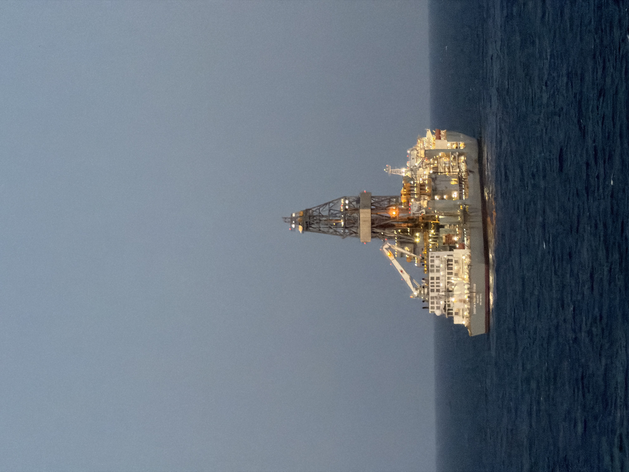 A nighttime shot shows an offshore drilling rig at sea. The rig is lit up with numerous lights. The background consists of a greyish-blue sky and the dark blue sea. There are no people visible. The location is not specified.