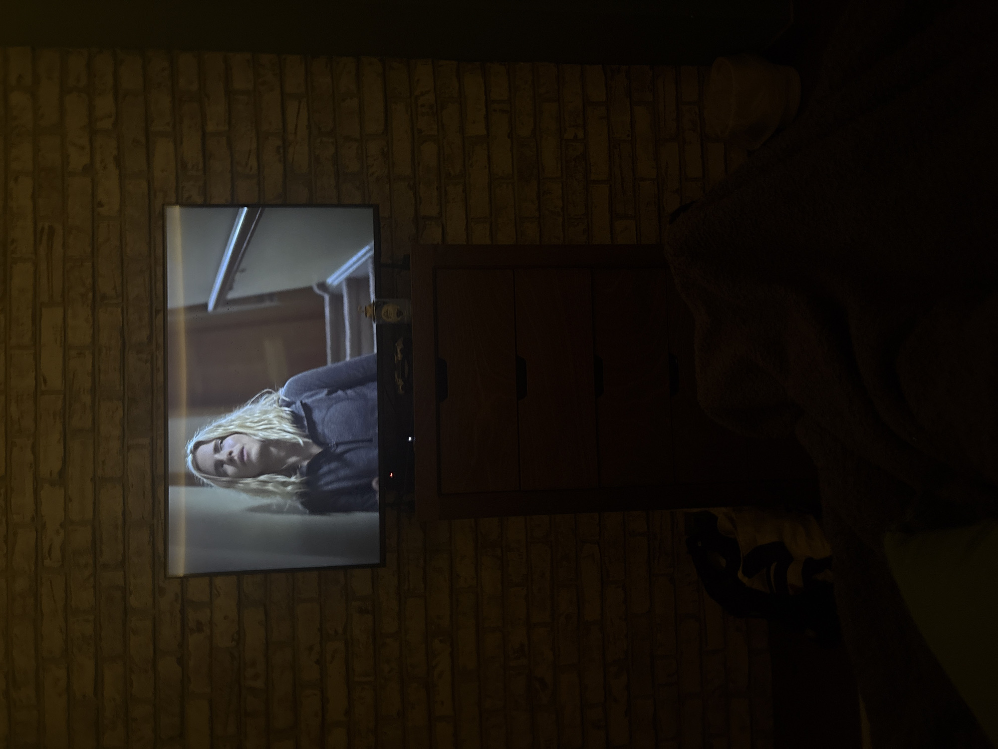The image depicts a room with a brick wall on which a television is mounted. The television screen displays a scene with a blonde woman lying down. Adjacent to the television is a dark-colored wooden cabinet with three drawers. To the right of the cabinet, there is a dark, amorphous shape that seems to be a couch or upholstered chair. The overall lighting in the room is dim, casting shadows and making details difficult to discern. The photograph was taken indoors. There is no information available in the image to confirm that the location of this photo is Huixquilucan de Degollado, Mexico.