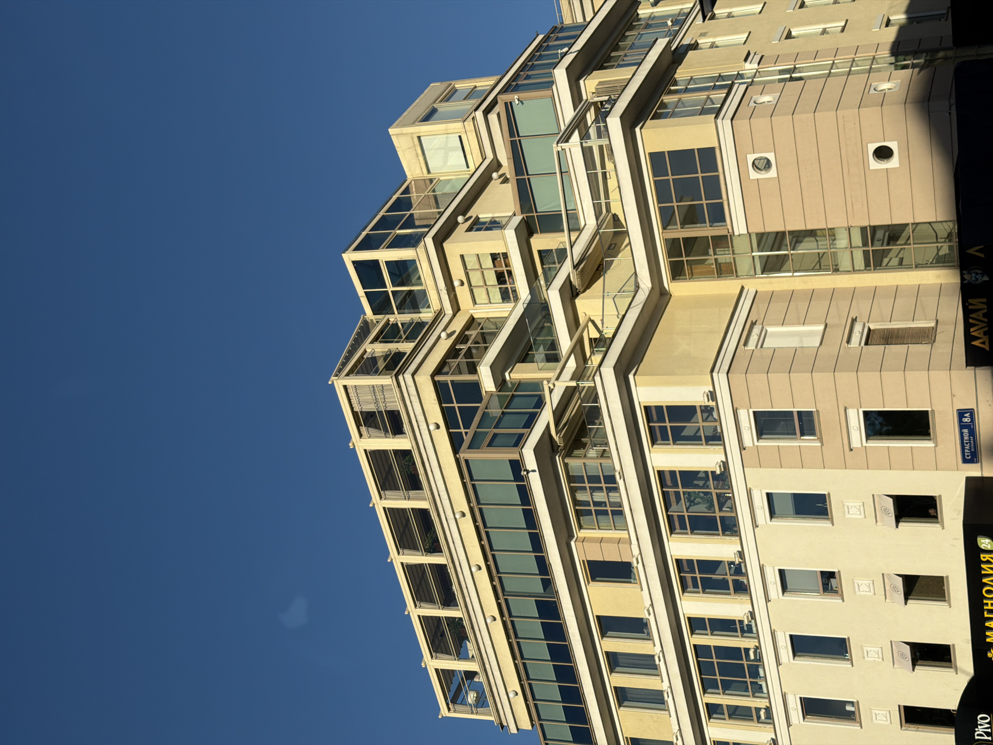 This is a low-angle, wide shot of a modern, multi-story building with a unique architectural design. The building is predominantly beige and cream-colored, featuring a staggered facade with numerous windows and balconies. Some balconies are enclosed with glass, while others are open, some with small plants visible. The building's structure appears angular and geometric, with recessed and protruding sections.
The sky is a clear, vibrant blue, suggesting a sunny day. The low angle of the shot emphasizes the height and scale of the building, with the vast expanse of the sky dominating the left side of the frame.
There is some visible text on the right side of the building. One sign, oriented vertically, reads "МАГНОЛИЯ 24" and below it, "PIVO". Another sign, oriented horizontally, reads "СТРАСТНОЙ 8А". These signs suggest the building may house businesses or apartments, and provide a potential street name and number.
The time of day appears to be late afternoon or early evening, indicated by the angle of the sunlight casting shadows and illuminating the building's facade. No people are clearly visible, though reflections in some windows hint at potential interior activity. The overall impression is of a contemporary urban environment in Moscow, Russia.
