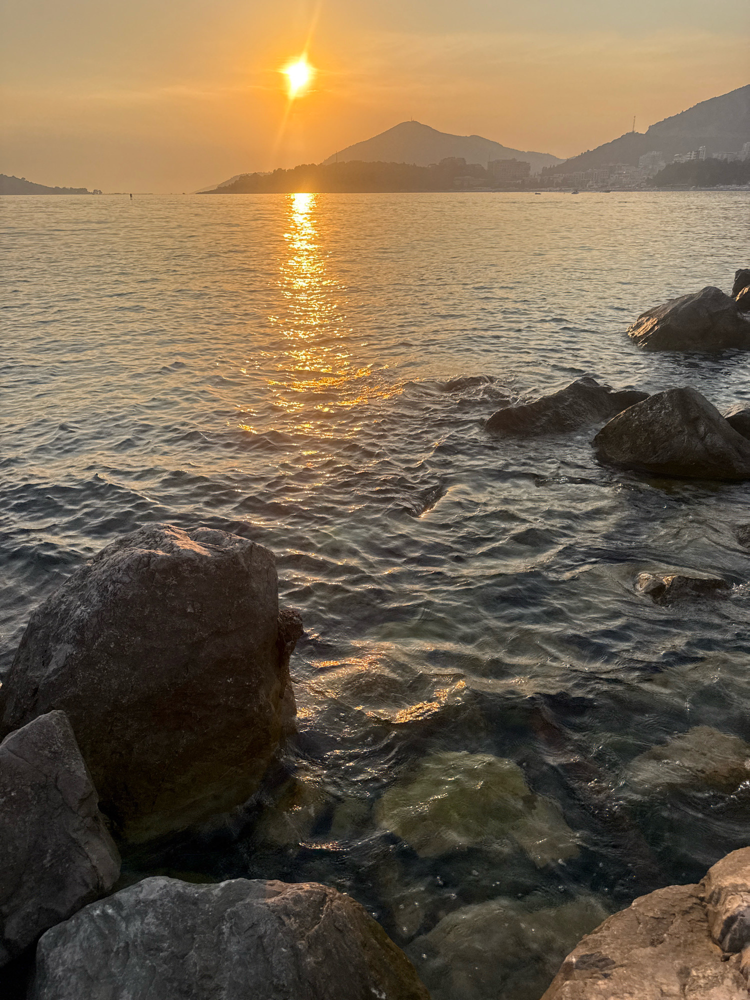 The image depicts a coastal scene in Rafailovići, Montenegro, during sunset. The sun, a bright orb, hangs low in the hazy, orange sky, casting a shimmering golden reflection across the calm water. In the distance, rolling hills dotted with buildings form the silhouette of the town. The foreground is occupied by large, dark rocks, some partially submerged in the clear water, others resting on the shoreline. The water is gently lapping against the rocks, creating small ripples. The overall atmosphere is serene and tranquil, characteristic of a Mediterranean evening.