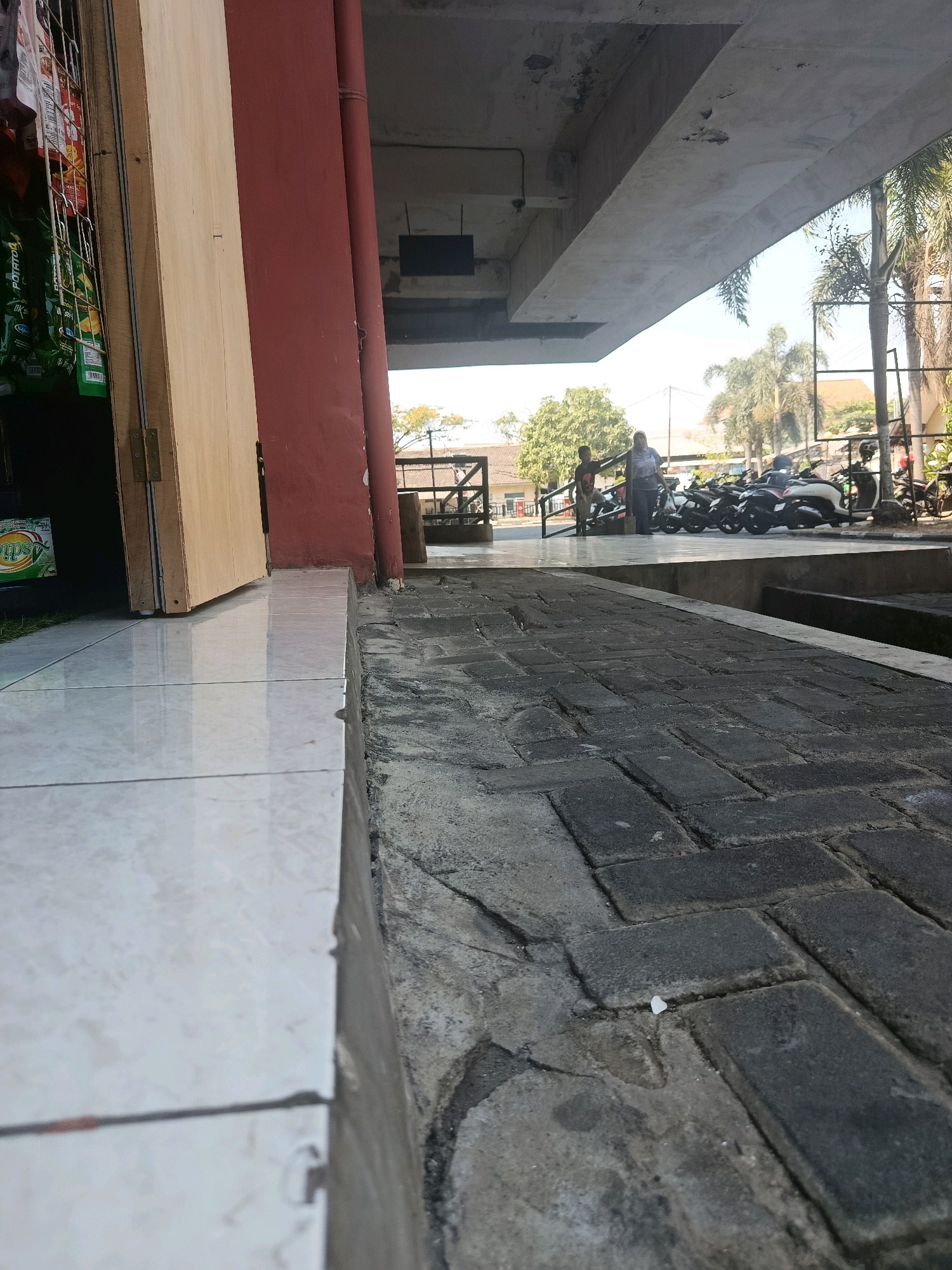 The image captures a low-angle view of a paved walkway and an adjacent building entrance in what appears to be a public area in Jobokuto, Indonesia. The walkway is constructed of dark rectangular bricks, showing some wear and tear, and is bordered by a white tiled edge. To the left, a wooden door is partially ajar, revealing shelves stocked with various bagged items, suggesting a small shop or stall. A red cylindrical pillar is prominent next to the door. In the background, under an elevated concrete structure, several motorcycles are parked, and two individuals are visible walking. Palm trees and other foliage are present, indicating a tropical or subtropical climate. The lighting suggests it is daytime.