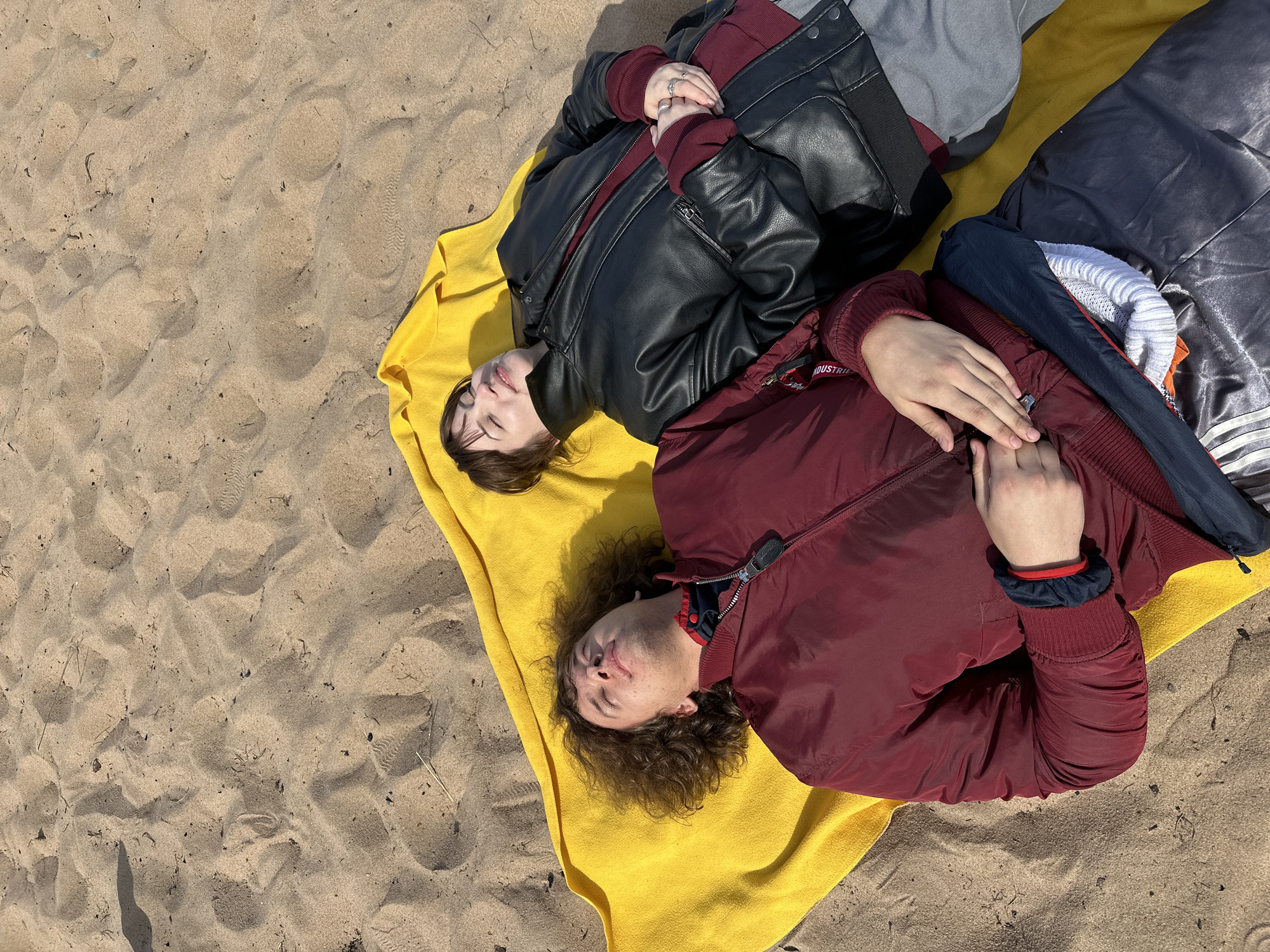 Two individuals are lying on a yellow towel on a sandy beach. The person on the left is wearing a black leather jacket over a maroon top and gray pants. The person on the right is wearing a maroon jacket. They both have their eyes closed. The location is Unknown, Unknown.