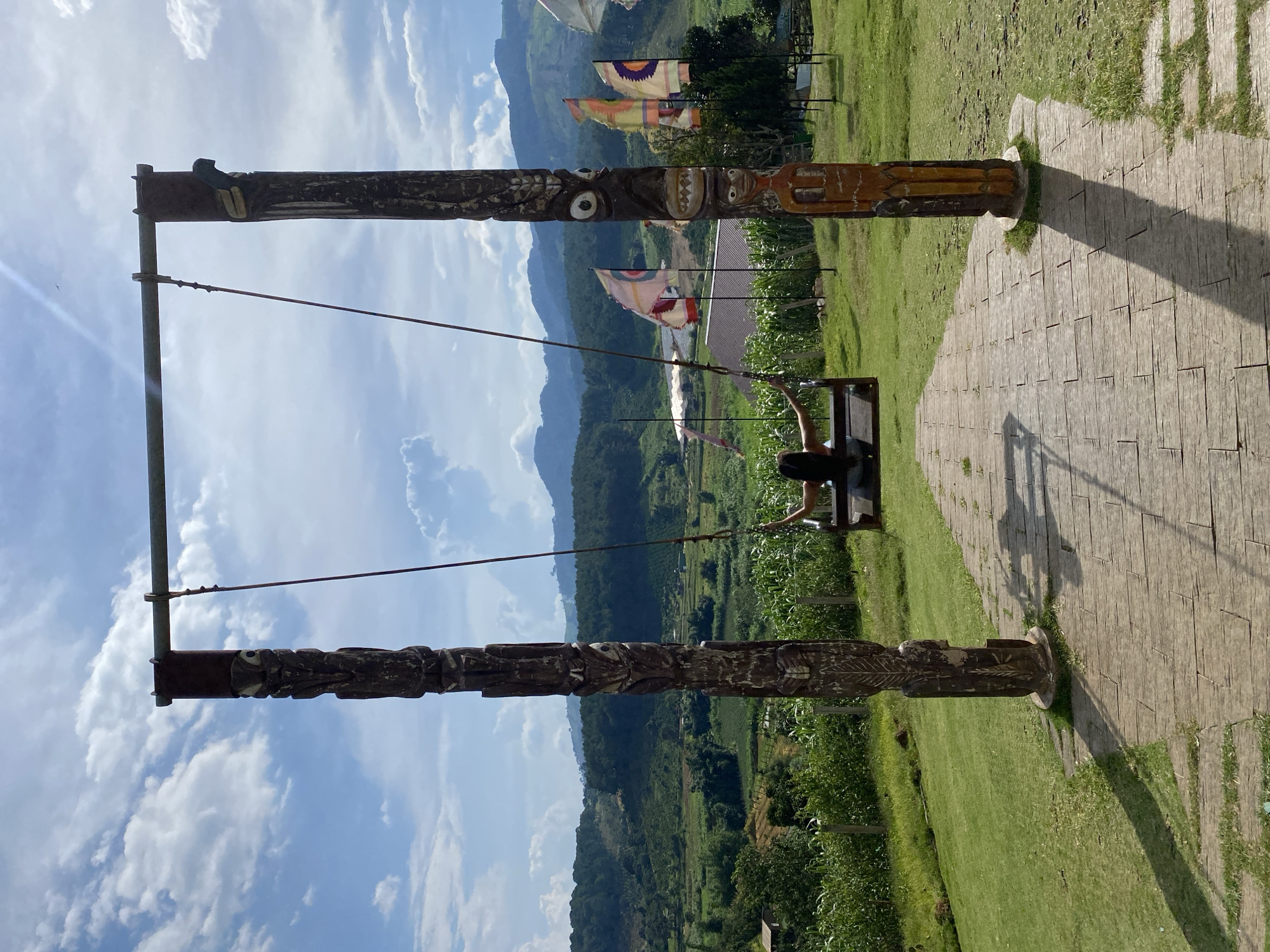 This media file captures a person on a swing with a scenic mountain backdrop. The swing set is constructed with two large, intricately carved wooden poles, each adorned with stylized faces and patterns. A person, with dark hair, is seated on the swing, which is suspended by ropes from a metal bar connecting the tops of the wooden poles.
The background reveals a lush green landscape with rolling mountains fading into the distance. Several colorful flags with geometric patterns are visible, adding a festive touch to the scene. The sky is a vibrant blue with fluffy white clouds, suggesting a pleasant day. The ground near the swing set is paved with rectangular stones, and to the right, a manicured grassy area is visible, casting long shadows from the swing structure. The overall impression is one of outdoor recreation and appreciation of natural beauty. The carved poles and flags hint at a cultural or tourist destination, likely in Vietnam, given the location context. The person on the swing appears to be enjoying the view and the gentle motion. The lighting suggests it is daytime, likely mid-morning or afternoon, with the sun casting distinct shadows. No visible text is present in the image.