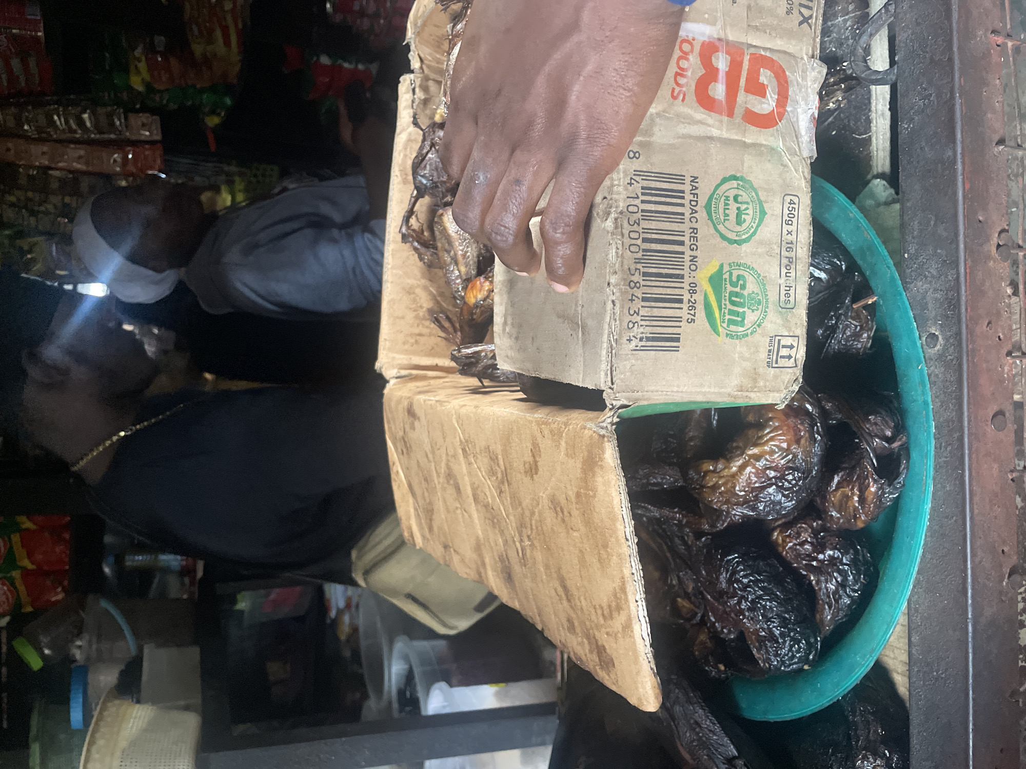 In Abuja, Nigeria, a close-up shot captures a person's hand resting on a cardboard box filled with dried fish. The box, displaying barcodes and certification labels for NAFDAC and Halal, is positioned in front of a green bowl containing more dried fish. In the background, out of focus, two individuals are visible, one wearing a black t-shirt and the other a light-colored headscarf and hoodie, suggesting a marketplace or food stall setting. The overall scene depicts a commercial transaction or display of food goods.