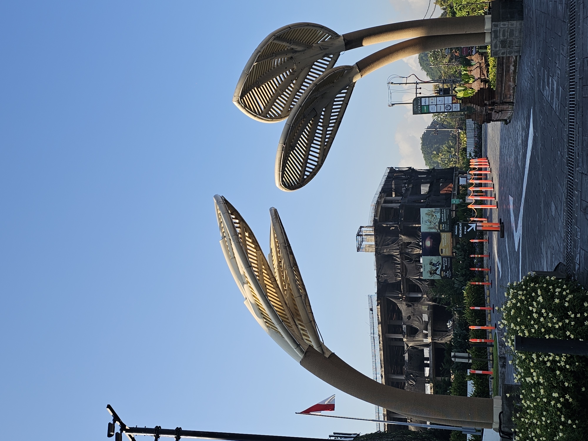 In Denpasar, Indonesia, an outdoor scene captures a bright, clear sky above a developing urban area. Prominently featured are two large, sculptural light fixtures shaped like stylized leaves, with textured, light-colored surfaces and ribbed interiors. To the left, a flagpole flies the Indonesian flag. In the background, a multi-story building under construction is covered in dark netting. Several signs are visible on and around the building, including one that reads "ICON BALI." The foreground shows a paved area with directional arrows, bordered by orange traffic cones and a low hedge of small white flowers. The overall atmosphere suggests a transitional or ongoing development project in a tropical urban environment.