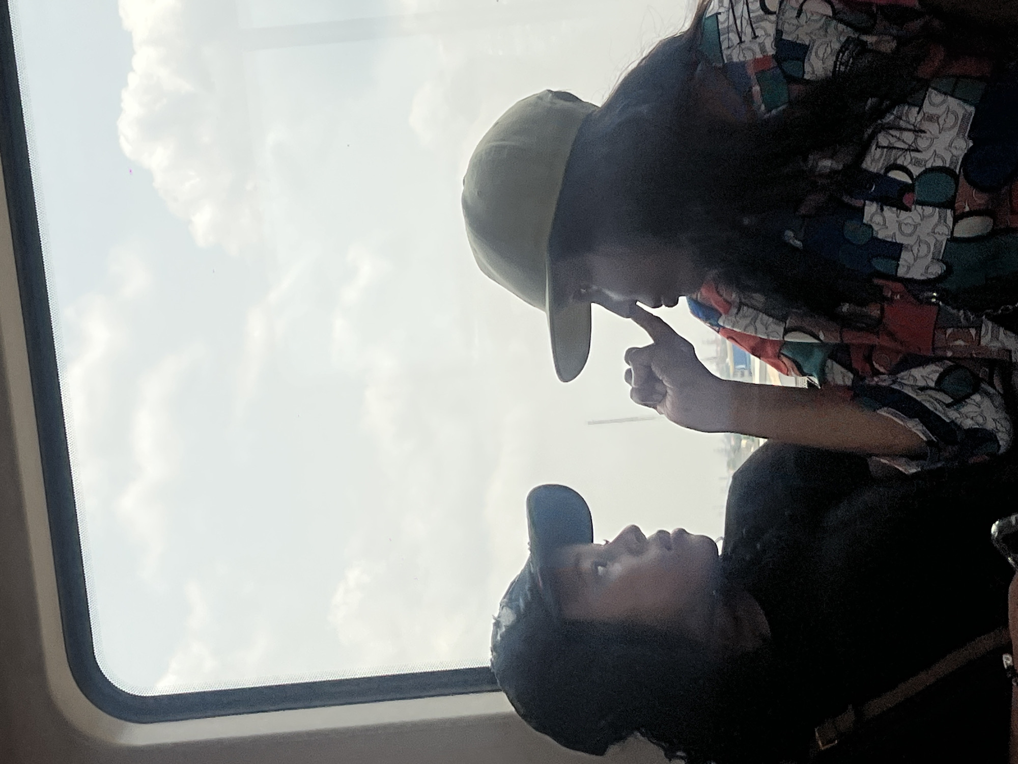 Two people stand next to a window, with clouds and a hint of buildings visible through the glass. One person, wearing a green cap and a patterned shirt with red, blue, and white elements, is extending their finger towards the other person's face, who is wearing a black cap and a black shirt. The window frame is visible on the left of the image. The location is Lagos, Nigeria.