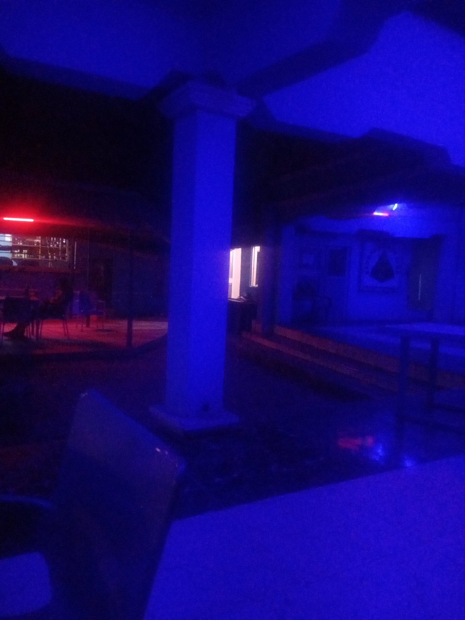 The image depicts an interior scene illuminated by predominantly blue and some red lighting, suggesting a nighttime or dimly lit environment. In the foreground, a dark, somewhat blurry object, possibly a piece of furniture like a table or chair, occupies the bottom portion of the frame.
To the left and mid-ground, red lighting illuminates a series of tables and chairs, implying a seating or dining area. Beyond this area, there is a structure with what appears to be a bar or counter, further suggesting a social or entertainment venue. A tall, white pillar is prominently visible in the mid-ground, extending upwards and seemingly supporting the ceiling structure.
In the background, a doorway or opening allows a view into another section of the establishment. This area is bathed in blue light, with some details of wall decor or fixtures visible. To the right, a raised area, possibly a stage or platform, is also illuminated by the blue lighting. The overall ambiance is that of a nightclub, bar, or similar entertainment space, likely located within the city of Sabon Gari Nassarawa, Nigeria. The scene appears to be unoccupied by people, or if present, they are indistinct and not a focal point of the image.