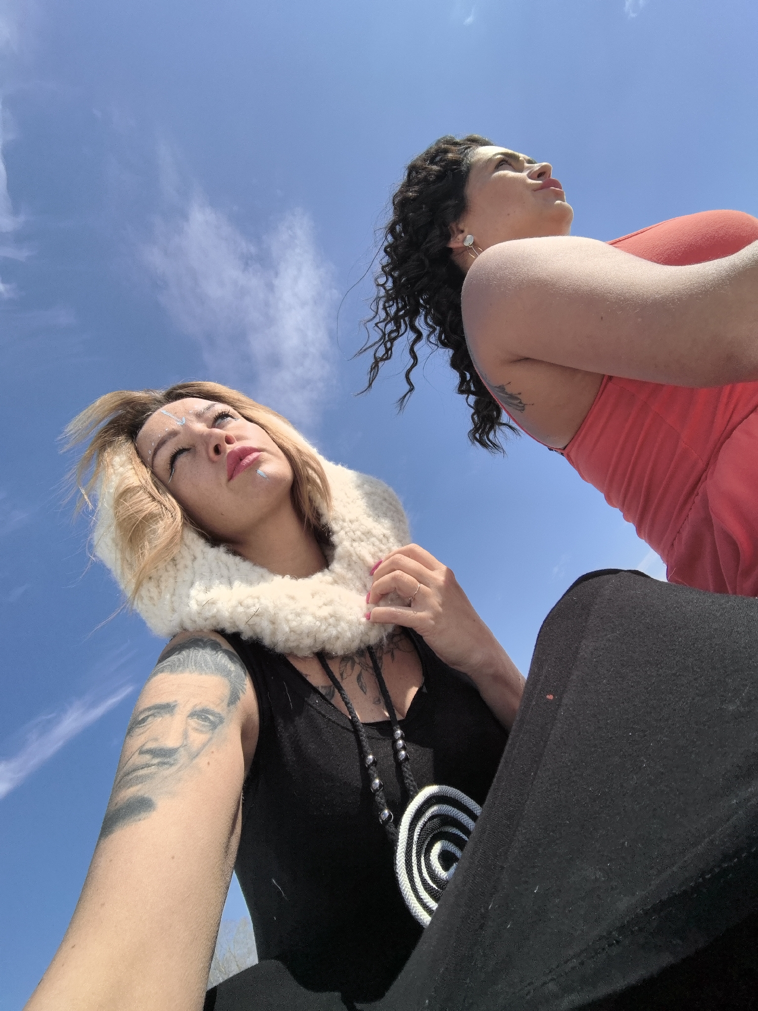 Two women are depicted from a low-angle perspective, looking upwards against a bright, clear blue sky with sparse, wispy white clouds. The scene is outdoors under strong daylight, potentially in Noviy, Russia, given the provided context.
In the left foreground, a woman with blonde hair wears a thick, white knitted neck warmer or cowl, a black top, and black pants. Her face features white or light blue dots and lines as decorative paint on her forehead and chin, and her lips are pink. She wears a prominent black and white spiral pendant necklace. A tattoo of a man's face is visible on her left upper arm, with other tattoos also appearing near her shoulder. She is seen holding her neck warmer.
To the right, partially visible in the background, is another woman with dark, curly hair. She is wearing a coral or reddish tank top or dress and is also gazing upwards. A tattoo is discernible on her upper arm. Both subjects maintain a contemplative or serene expression as they look towards the sky. No visible text is present in the image.