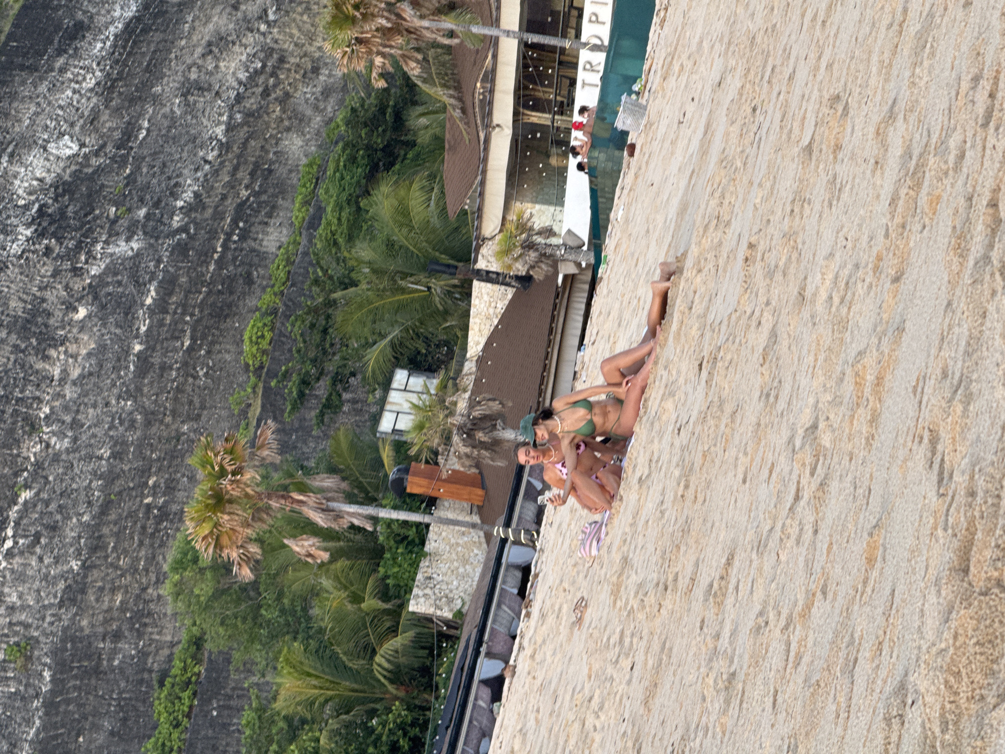 The image captures a vibrant tropical scene in Kuta Selatan, Indonesia, depicting a sandy leisure area adjacent to a resort and a dramatic cliff face. In the foreground, two women are relaxing on the light-colored sand, appearing to sunbathe or lounge. One woman wears a green bikini and a green hat, accessorized with a necklace. The other wears a pink and white patterned bikini. A striped item, possibly a towel or bag, is visible near them.
Behind the women, the scene transitions to a resort-like complex. Buildings with brown roofs and modern architecture are visible, alongside what appears to be a swimming pool or a section of the ocean. Several other people are seen near the water and buildings, enjoying the facilities. The word "TROPI" is distinctly visible on the facade of one of the buildings, likely part of a name such as "Tropical" or "Tropicana."
Further in the background, a large, rugged, dark-grey cliff face dominates, characterized by its textured surface and white streaks. Portions of the cliff are covered with lush green vegetation and numerous palm trees, contributing to the tropical ambiance.
The lighting suggests it is daytime, possibly late afternoon, with a warm glow over the scene. The weather appears clear and sunny, enhancing the leisurely and relaxing atmosphere of this Indonesian coastal location.