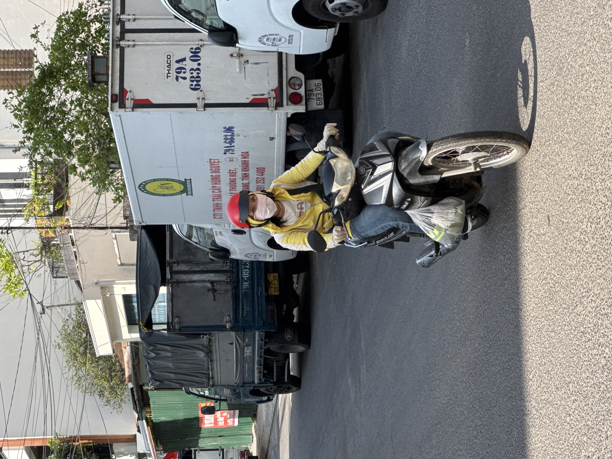 A woman wearing a yellow jacket, blue jeans, a red helmet, and a medical mask is riding a dark-colored scooter on a paved street in Nha Trang, Vietnam. She is carrying a clear plastic bag filled with items on the front of the scooter. The scooter's front wheel casts a circular shadow on the road. Behind her, there are several trucks. The truck closest to the viewer is white with blue lettering that includes the text "CTY TINH TRÁI CÂY HUNG NGUYET" and "79A 683.06". Further back, a blue truck with a tarp-covered bed is visible, and on its rear bumper, there is a yellow license plate that reads "79C 097.35". Another white truck is partially visible to the right of the first white truck. The background consists of buildings and trees under a clear sky.