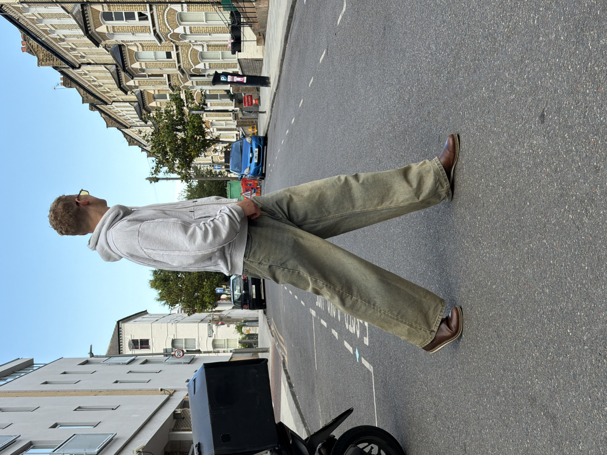 A young white man with blond hair and glasses is walking across a street in London, United Kingdom. He is wearing a light gray hooded sweatshirt and loose-fitting, light brown or olive green jeans. He has on brown leather loafers. He is walking from the left side of the frame towards the right. In the background, there is a street lined with terraced houses. A blue car is parked on the right side of the street, and a black car is parked further down on the left. A motorcycle with a large black box on the back is on the left side of the street. The sky is clear and blue. There are traffic signs and streetlights visible.