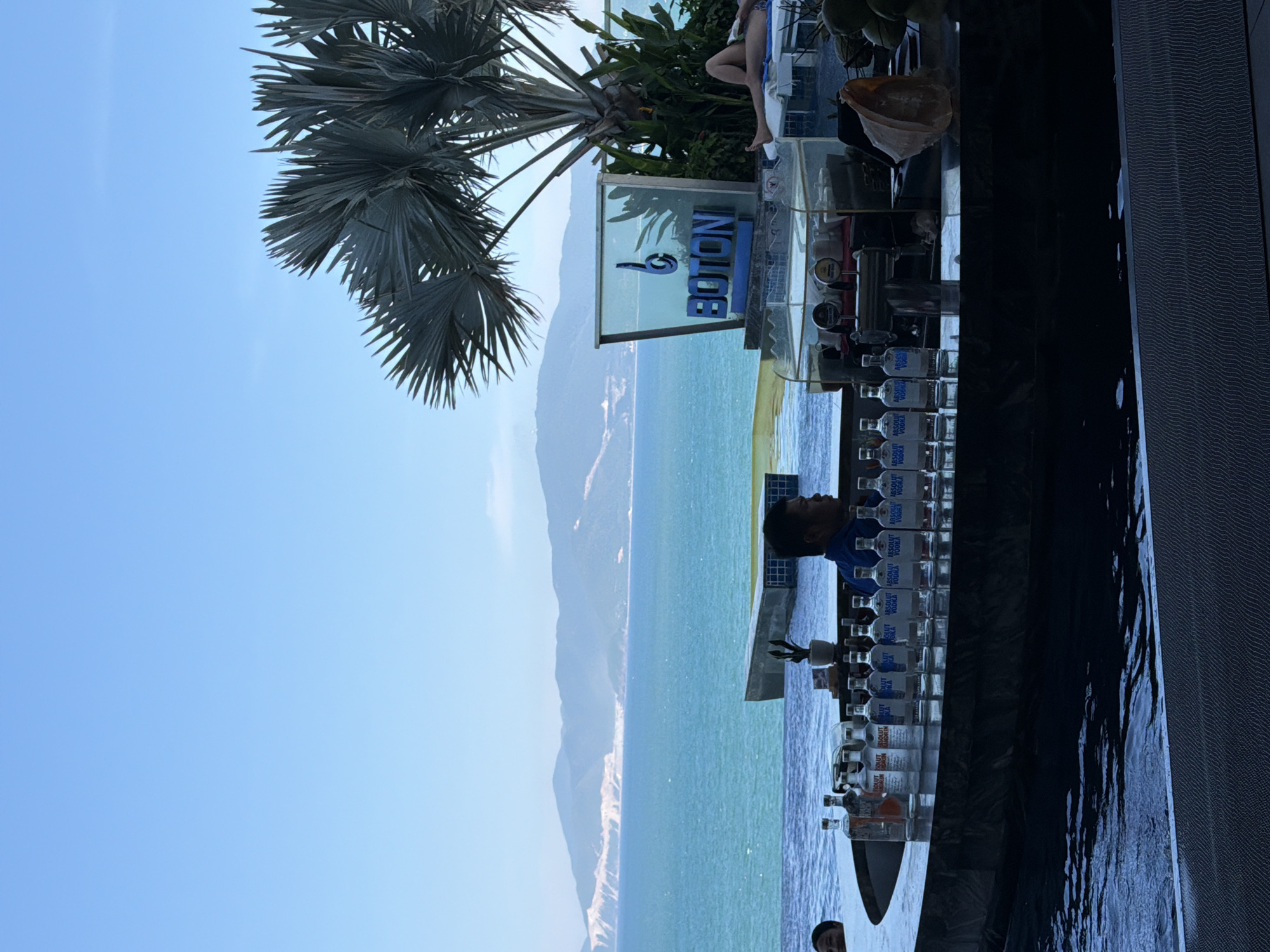 The image shows a bar area overlooking the sea in Bac Nha Trang Ward, Vietnam. A bartender, a man wearing a blue shirt, stands behind a counter filled with bottles of Absolut vodka and other alcoholic beverages. In the background, a swimming pool and palm trees are visible. A sign with the word "BOTON" is illuminated. The sea is turquoise, and mountains are visible in the distance. The sun is shining, and the sky is clear blue.
