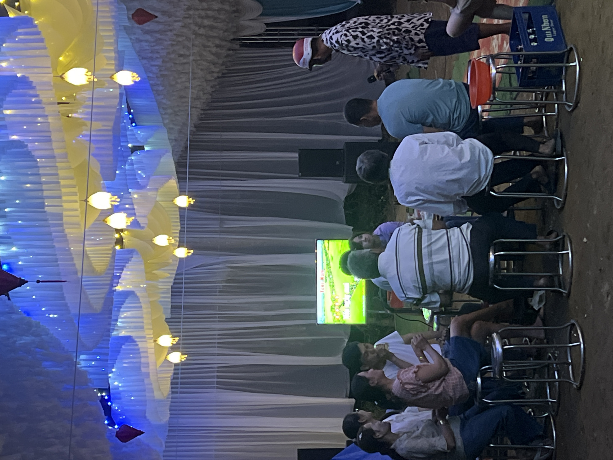 This image depicts a nighttime or evening social gathering in what appears to be an outdoor or semi-outdoor setting, possibly under a large tent or canopy. The overhead structure is elaborately decorated with undulating blue and yellow/gold fabric panels, illuminated by numerous strings of fairy lights in matching colors.
Several individuals are present, predominantly adults, mostly seated on simple metal stools. Their attention is directed towards a central flat-screen television displaying a brightly colored animated scene, which could be a cartoon or a karaoke display. One person, wearing a patterned shirt and a red and white helmet, stands near the back, holding a microphone, suggesting singing or announcements are part of the event.
Other visible objects include a red bowl resting on one of the stools, and a blue plastic crate clearly labeled "Qui Nhon Bia Lager," a brand of beer from Vietnam, supporting the provided location context of Bình An, Vietnam. Speakers are also visible in the background. The ground beneath the seating area appears to be unpaved earth. The overall ambiance suggests a casual, communal event focused on entertainment and socializing. The specific emotions of the attendees are not clearly discernible due to the distance and angle.