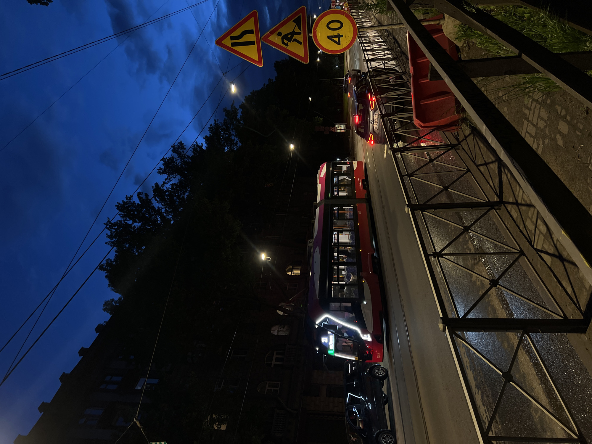 This is a nighttime street scene in Saint Petersburg, Russia, with a modern tram in motion. The tram is predominantly red and white, with illuminated blue and white accents along its sides and front. It is traveling on tracks on a wet asphalt road, reflecting the streetlights and tram's own illumination. Several cars are also visible on the road, one a black SUV closely following the tram, and another, a dark sedan with illuminated taillights, further down the street.
The street is lined with a metal fence, separating the roadway from a sidewalk. Beyond the fence, a red construction barrier is visible. The background features a dark, multi-story building with arched windows, characteristic of Saint Petersburg's architecture. Tall trees with dark foliage overhang the building and the street, contributing to the nighttime ambiance. The sky is a deep blue with hints of clouds, indicating either twilight or a very dark night.
Traffic signs are present on the right side of the image. A triangular yellow sign with a black arrow indicating a narrowing road is stacked above another triangular yellow sign depicting a worker with a shovel, signaling road work. Below these, a circular yellow sign with a black border displays the number "40", indicating a speed limit of 40 kilometers per hour. Overhead, power lines crisscross the sky. The overall impression is an urban street scene at night, with the city's infrastructure and a functioning tram system evident.