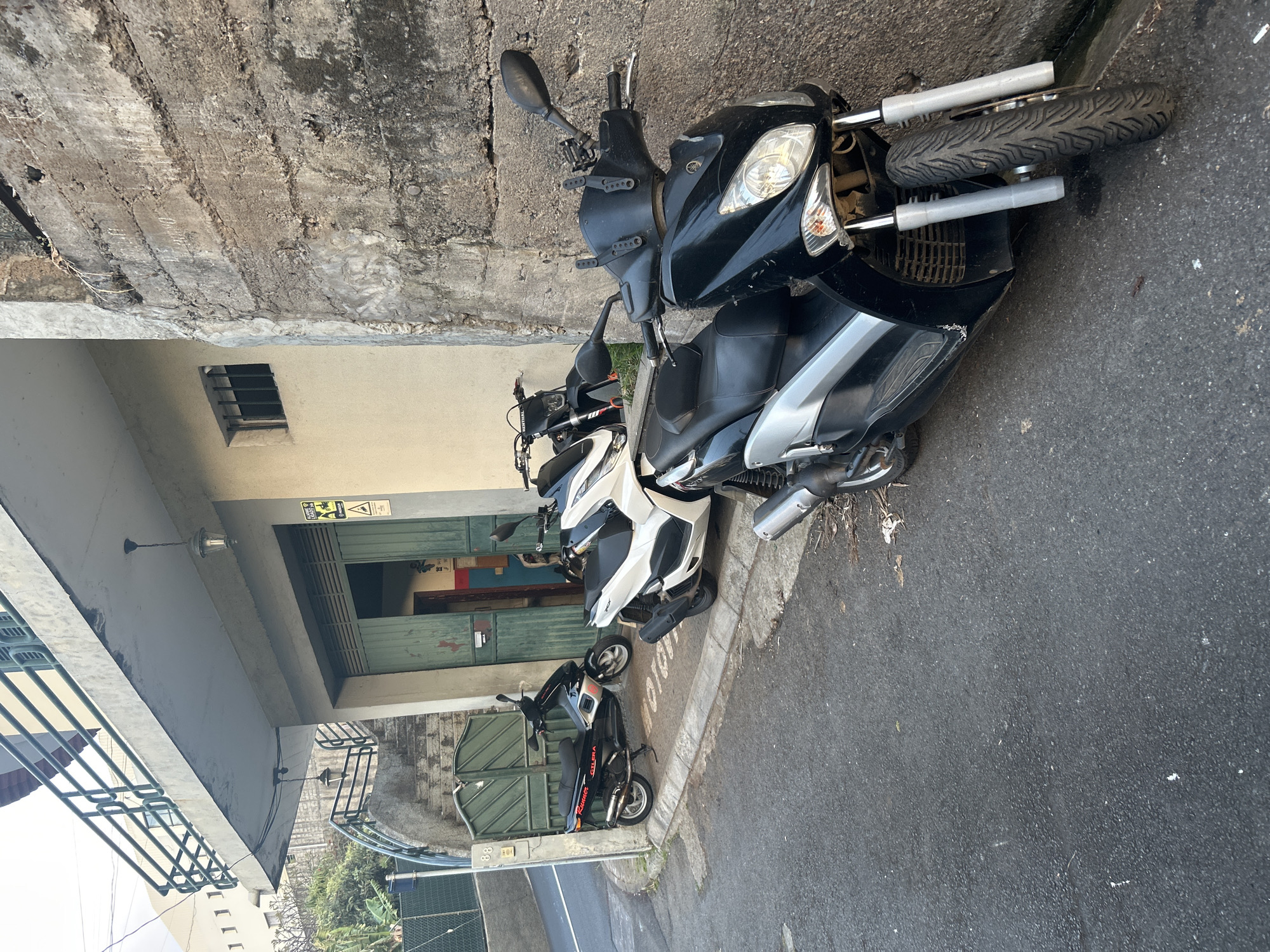 The image depicts a street scene in Câmara de Lobos, Portugal, featuring a row of parked scooters. Three scooters are visible: one black, one white, and one dark-colored scooter parked behind the others. They are parked on the side of the road next to a building with a concrete exterior. A barred window is visible on the building. The scooters are parked on the asphalt road. The lighting appears to be natural daylight.
