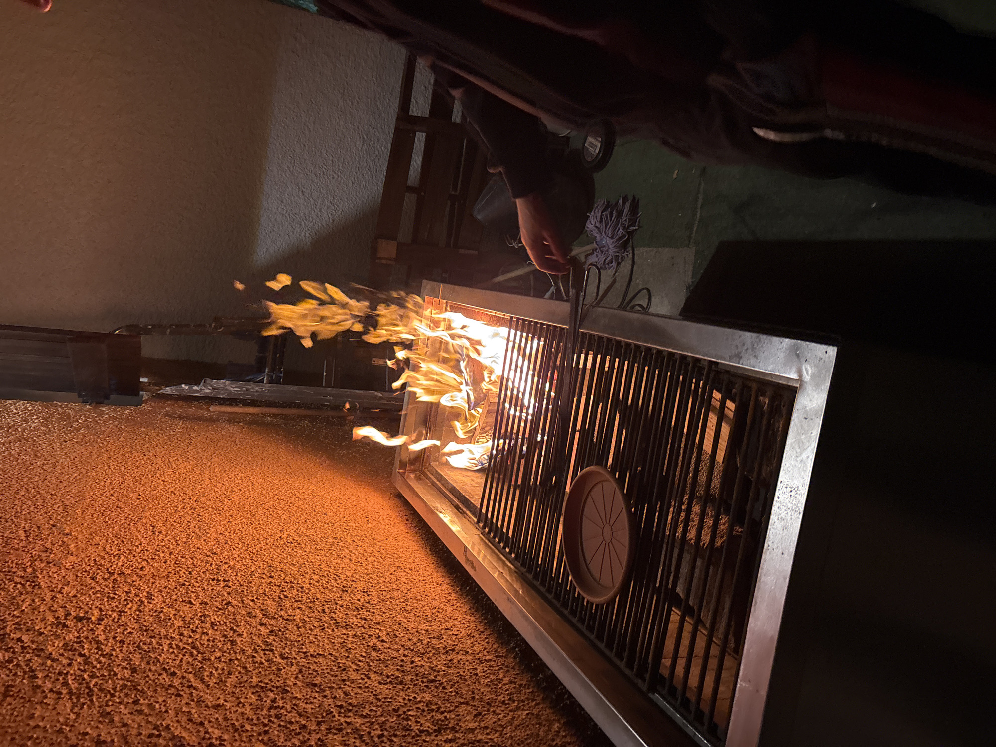 A person is tending to a fire in a grill in Vila Real, Portugal. The grill is a rectangular metal structure with vertical bars, and a round object affixed to the side. Flames are rising from the grill's interior. The ground is orange. A person's hand is visible near the top of the grill. Part of the person's clothing is visible at the top. The environment includes a wall, a structure, and a green surface in the background.
