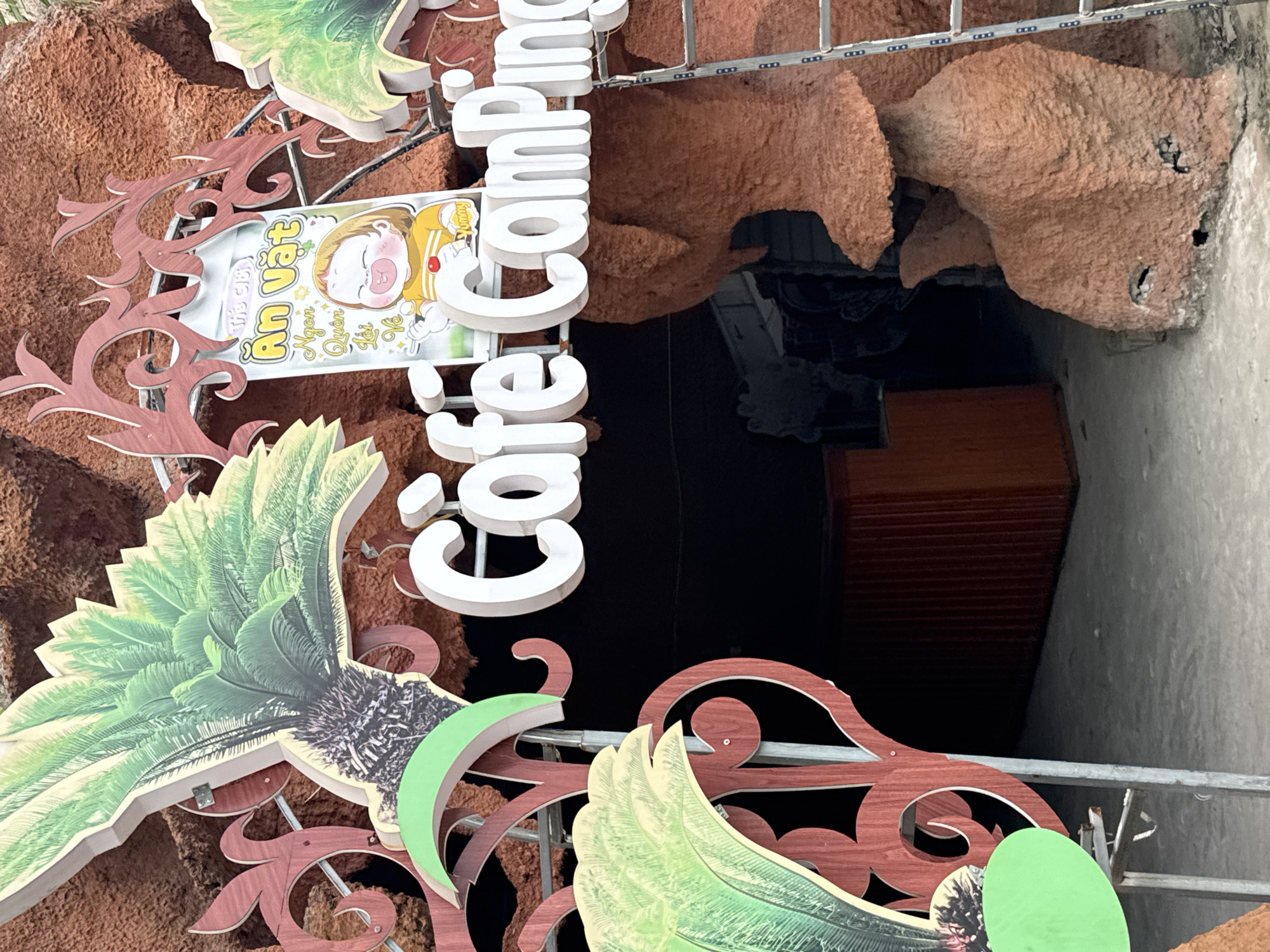This is a photograph of the entrance to a cafe and camping establishment in Hoa Lư Ward, Vietnam. The entrance features a large, rustic, reddish-brown, artificial rock formation. A prominent sign above the entrance reads "Cafe Camping" in large, white, 3D lettering. To the left and right of the sign are decorative elements resembling palm fronds, rendered in green and yellow. Between the palm fronds and the "Cafe Camping" sign, a smaller sign depicts a cartoon drawing of a child eating, with Vietnamese text that appears to read "An Vặt Ngon Quen Tôi" (Delicious Snacks I'm Familiar With). A silver ladder leans against the right side of the rock formation. In the background, a dark opening suggests the interior of the establishment, with a glimpse of what looks like a wooden counter. The ground in front of the entrance is paved.