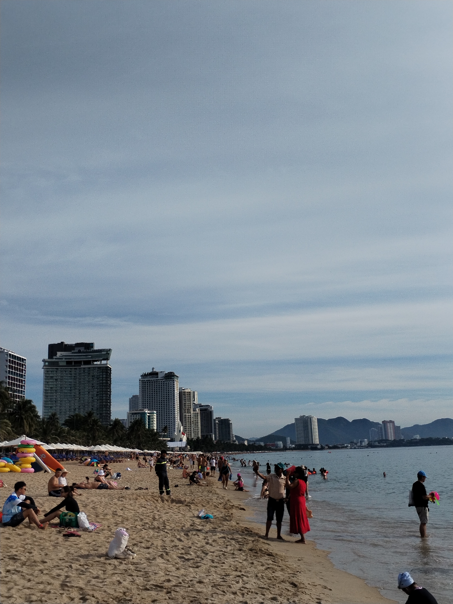 This is a vertical, eye-level shot of a crowded beach scene in Nha Trang, Vietnam. The foreground is dominated by a sandy beach with several people scattered across it. Some are sitting or lying down, while others are standing. In the middle ground, the ocean is visible with people swimming and wading. The background features a cityscape with numerous tall buildings and a mountain range in the distance. The sky is partly cloudy.
Specifically, on the beach, several individuals are visible in various states of relaxation and activity. Near the left side of the image, two men are seated on the sand, one wearing a blue and white shirt and shorts, and the other in a black shirt and shorts. Nearby, a woman in a black top and shorts is also seated. Further back, a group of people are gathered, some of whom appear to be sunbathing. In the middle of the beach, a man in dark clothing and a reflective vest stands, seemingly observing the beachgoers. Two individuals, one in a black shirt and the other in a red dress, are interacting near the water's edge.
The water itself contains numerous people, with some swimming and others standing closer to the shore. A man in a blue cap is standing in the water on the right side of the image, holding a colorful object.
The cityscape in the background consists of modern high-rise buildings, characteristic of a coastal tourist destination like Nha Trang. The presence of umbrellas and beach amenities suggests a popular recreational area. The overall atmosphere appears to be one of a busy, sunny day at the beach.