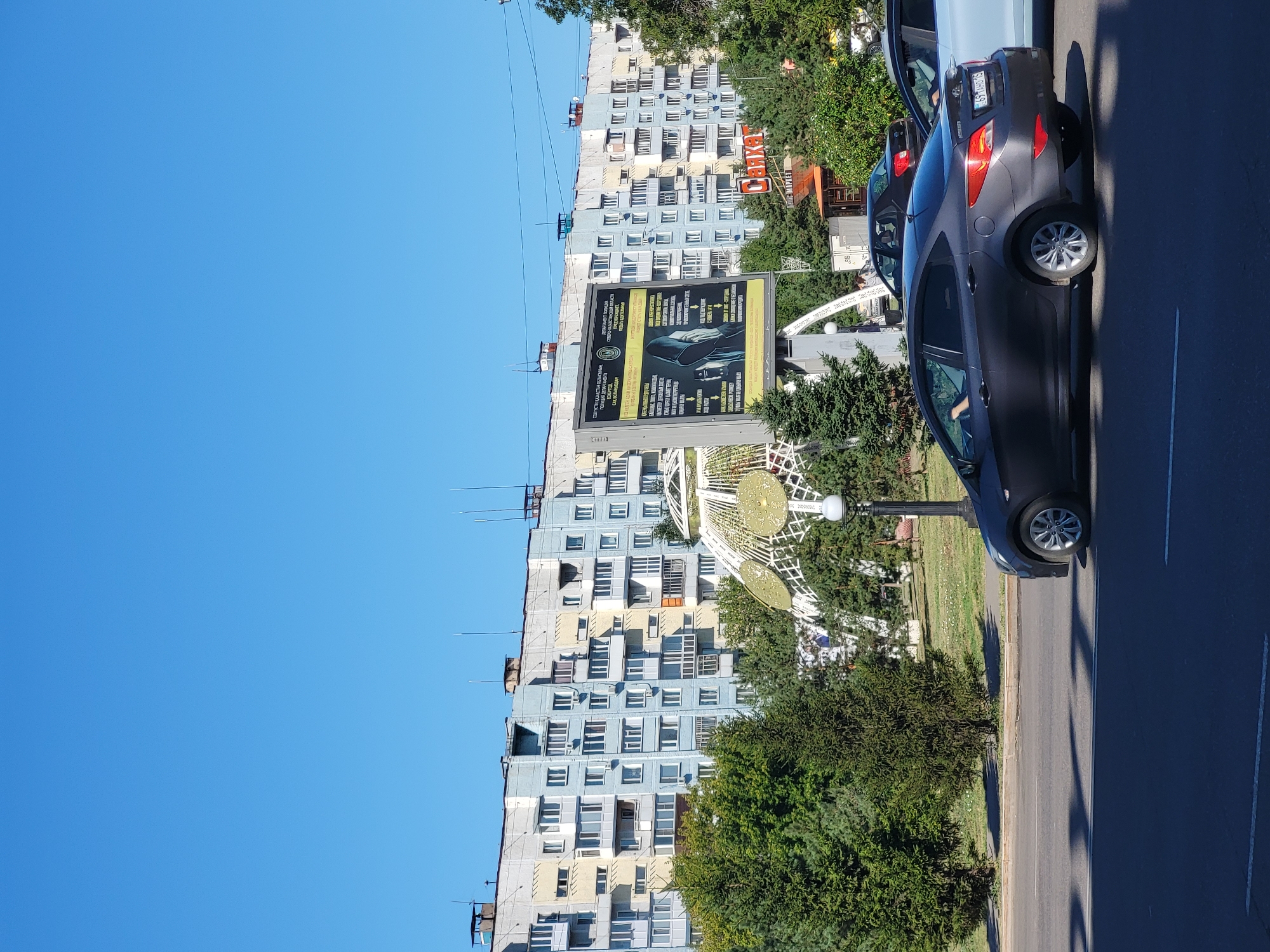 An eye-level, outdoor, medium shot captures a sunny day in Petropavl, Kazakhstan, featuring a long, multi-story apartment building with a pale blue and yellow facade. The building has numerous balconies and windows, with several antennas on its roof. In front of the building, a large digital billboard displays text in Cyrillic and an image of a person wearing a hooded jacket, with other graphics and text. To the left of the billboard is a decorative, dome-like structure with a white lattice and golden accents. A black sedan is in the foreground, driving on a paved road. Trees and greenery are visible between the billboard and the apartment building, as well as to the right of the scene. A street lamp is positioned near the decorative structure. The sky is clear and blue.